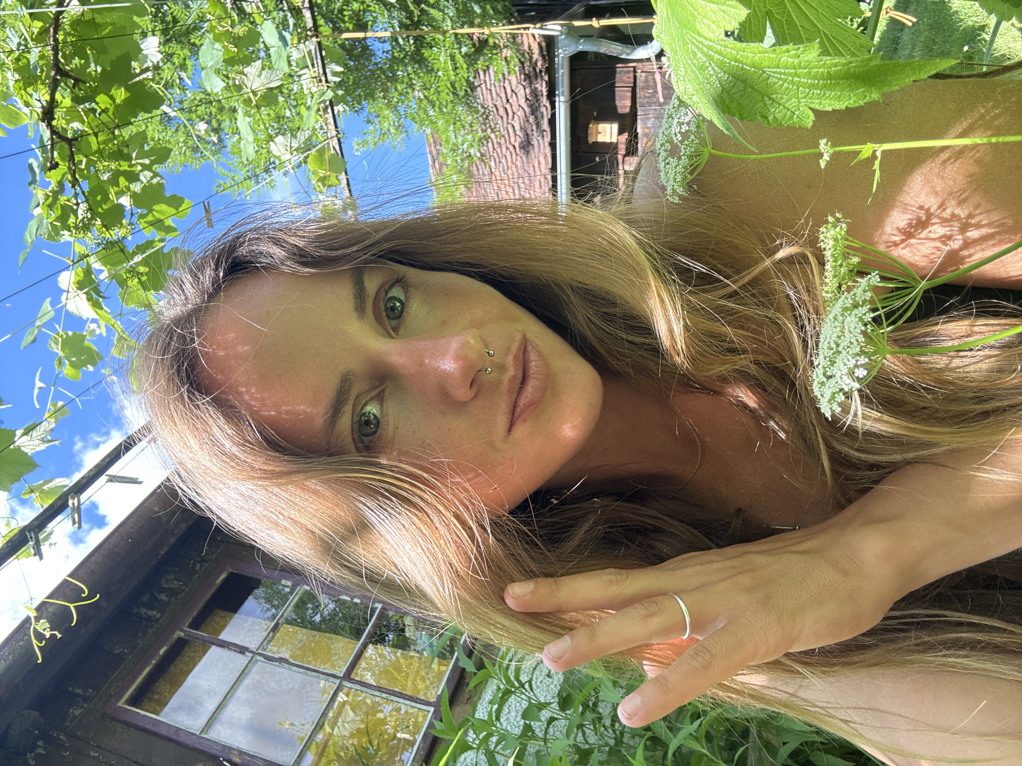 An outdoor, medium-shot, selfie-style photograph shows a woman with long, blonde hair and green eyes, wearing a silver nose ring and a silver ring on her left index finger. She is in a garden setting, with green foliage and a rustic building in the background. The woman is looking directly at the camera, with her left hand held up near her face, obscuring part of her forehead. The lighting suggests it is daytime, with sunlight casting shadows on the scene.