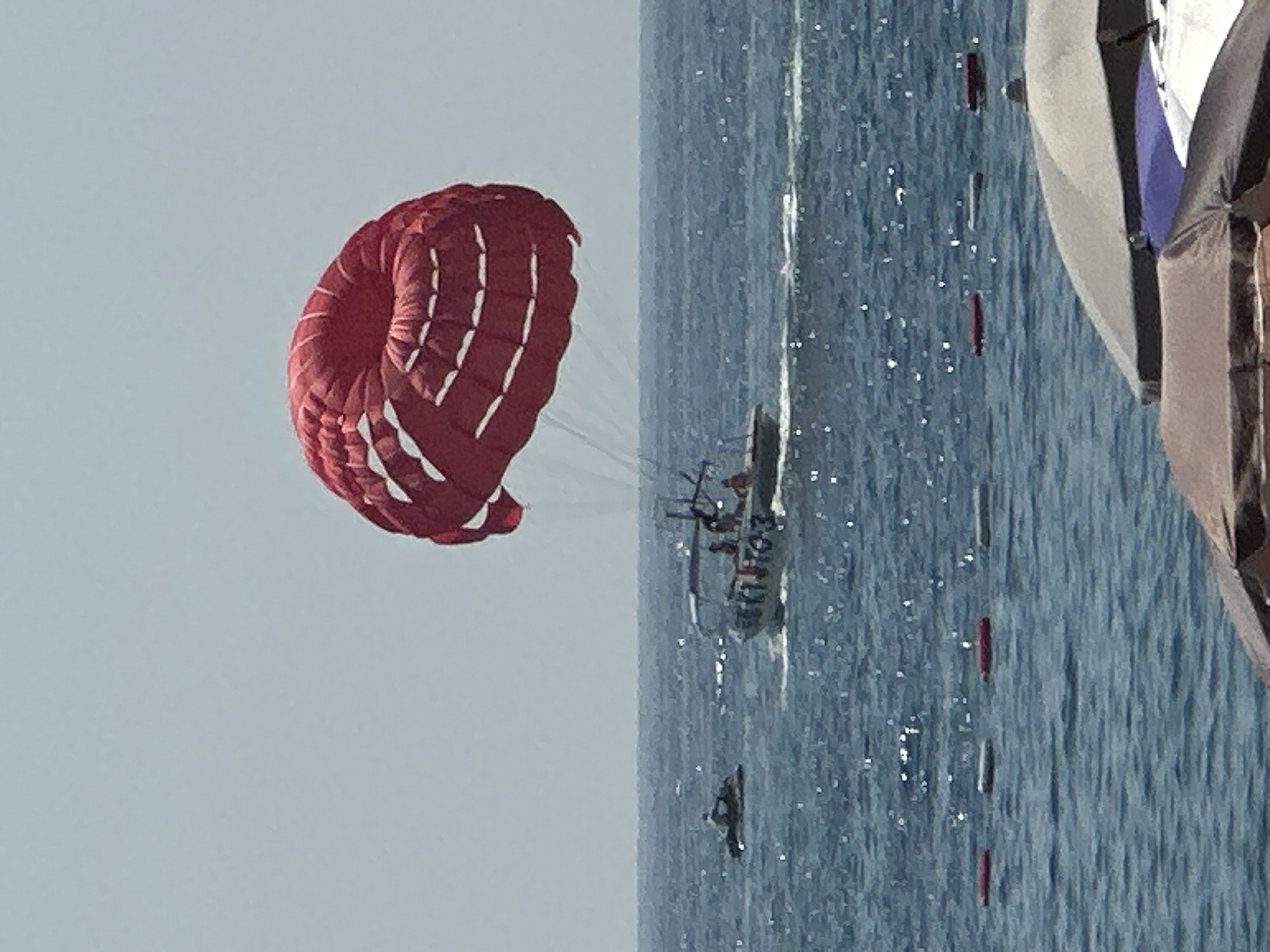 The image depicts a sunny day at the sea, likely in Budva, Montenegro, given the parasailing activity and beach umbrellas. In the foreground, several beach umbrellas are visible, suggesting a beach setting. The middle ground shows the blue, choppy sea with sunlight glinting off the surface. A boat, identified by the numbers "503", is actively engaged in parasailing. A red parachute, carrying unseen individuals, is airborne, towed by the boat. To the left of the boat, a jet ski is also present on the water. The background consists of a clear, pale blue sky. The overall scene captures a moment of recreational activity on the water.