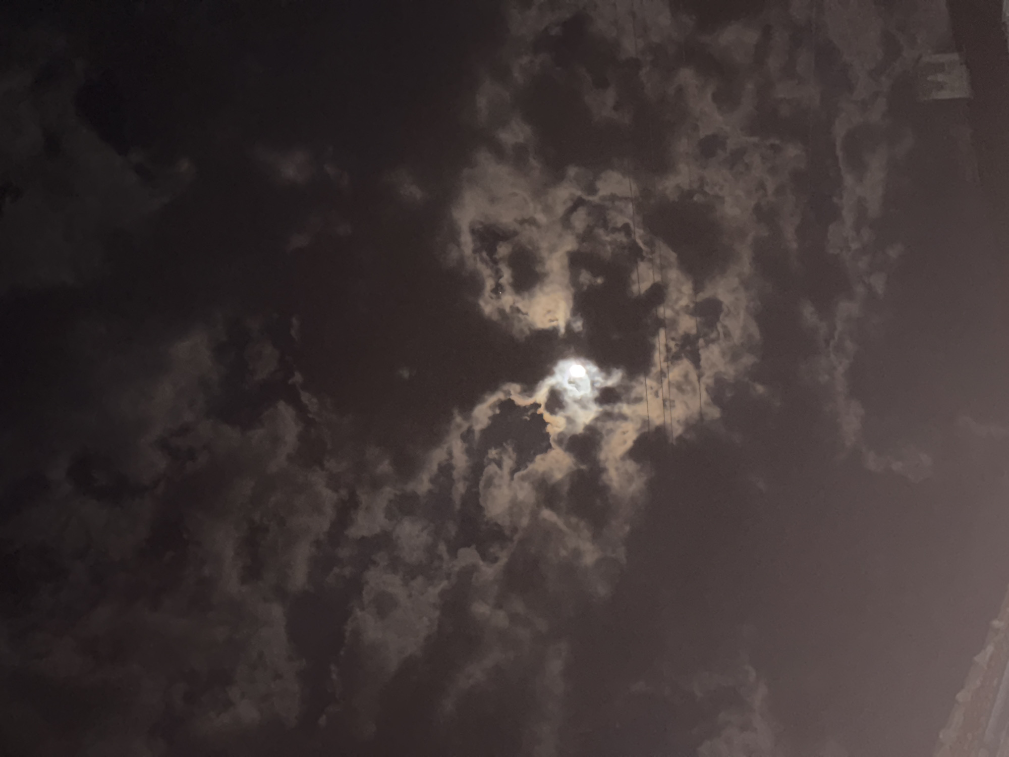 The image shows a night sky filled with dark, dense clouds. A bright moon is partially obscured by the clouds, with its light filtering through the gaps. There are some powerlines visible in the top right quadrant, running vertically through the image. The darkness and cloud cover suggest an overcast night, with only the moon providing any significant illumination. The photo appears to be taken in Sarnevo, Bulgaria.