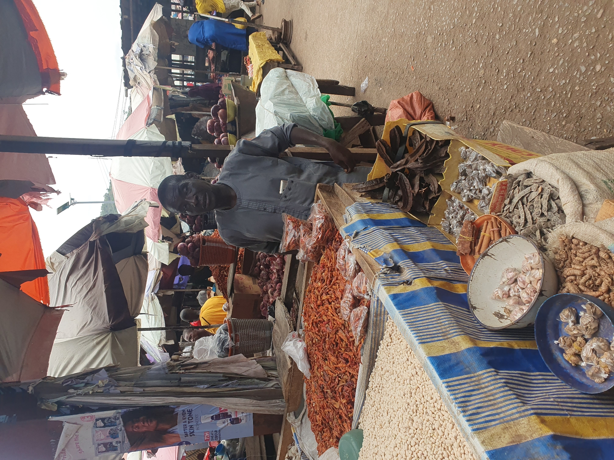 The image shows a man seated behind a market stall in Lokoja, Nigeria. He is wearing a gray shirt and is positioned behind a table laden with various food items and spices. The table is covered with a blue and yellow striped cloth. Items on display include dried peppers, beans, spices in bags and small dishes, and other unidentified goods.
In the background, there are more market stalls under tarps and umbrellas, with other vendors and customers visible. The ground appears to be paved. A poster for a skin-lightening product is visible among the market clutter.