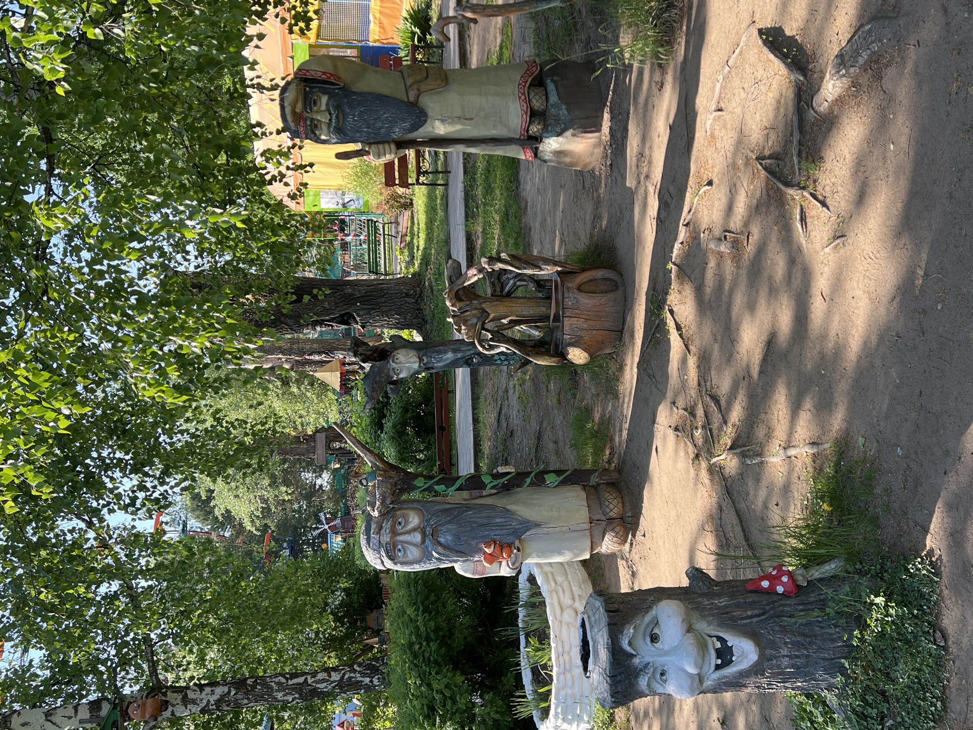 The image depicts a row of carved wooden sculptures in a park or recreational area. The location is identified as Chaikovsky, Russia.
The sculptures appear to represent folklore or fairytale characters. The figures include depictions of bearded men with distinct headwear and clothing details, as well as more abstract or fantastical creations.
The sculptures are positioned along a dirt path or walkway, with vegetation and trees visible in the background. The area seems to be well-maintained with evidence of other recreational amenities. The lighting suggests a sunny day.