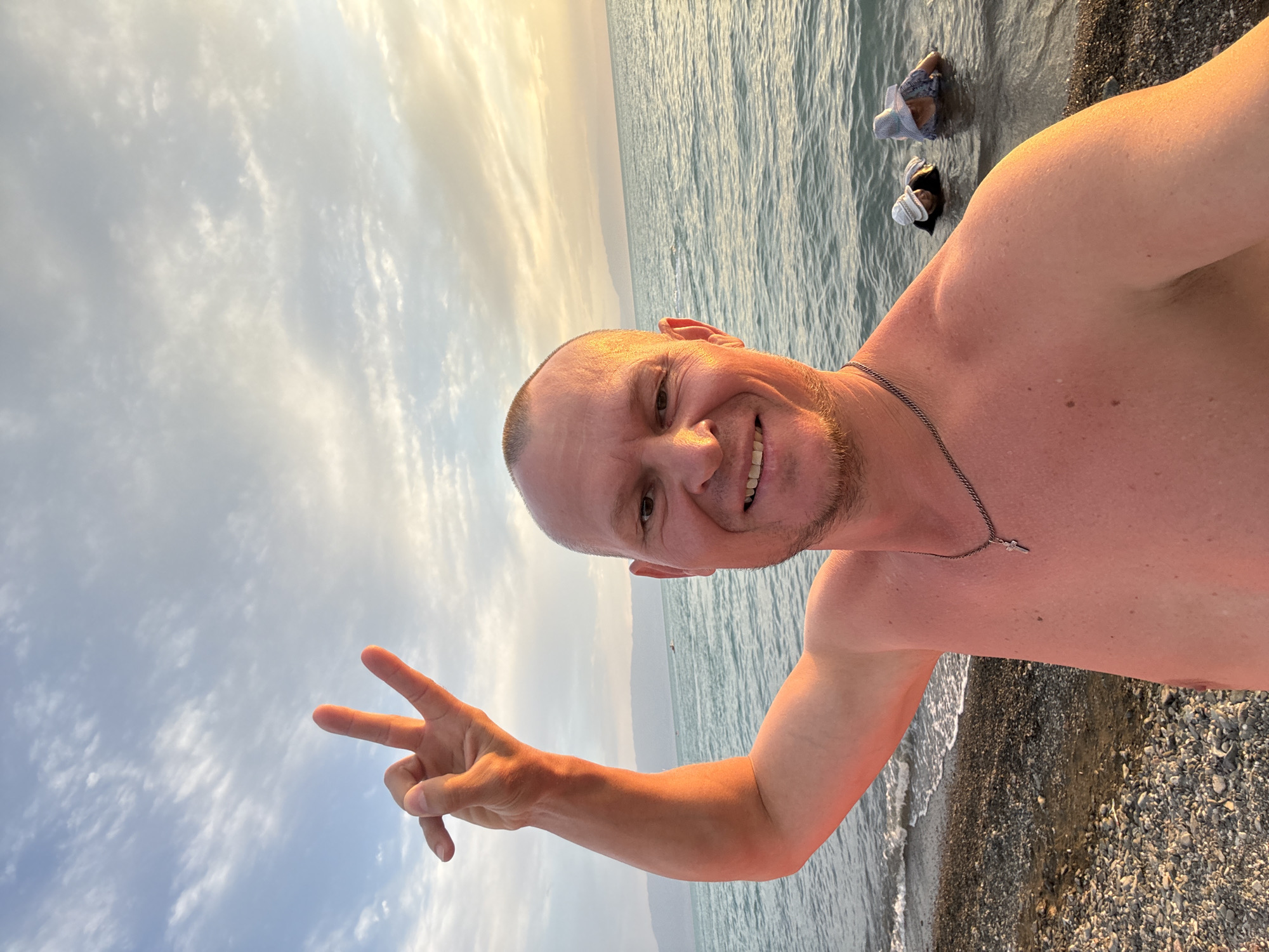 A selfie taken by a shirtless bald man standing on a pebble beach. He is smiling and making a peace sign with his left hand, which is raised. The man is wearing a silver chain with a cross pendant. Behind him, there is a body of water, likely a lake or sea, with gentle waves lapping the shore. In the water, two people, possibly children, are wearing hats and appear to be wading or swimming. The sky is cloudy and illuminated by the warm light of the setting sun, casting an orange glow on the scene. The background shows a distant mountain range under a hazy sky. The location is specified as the city of Urdzhar, Kazakhstan.