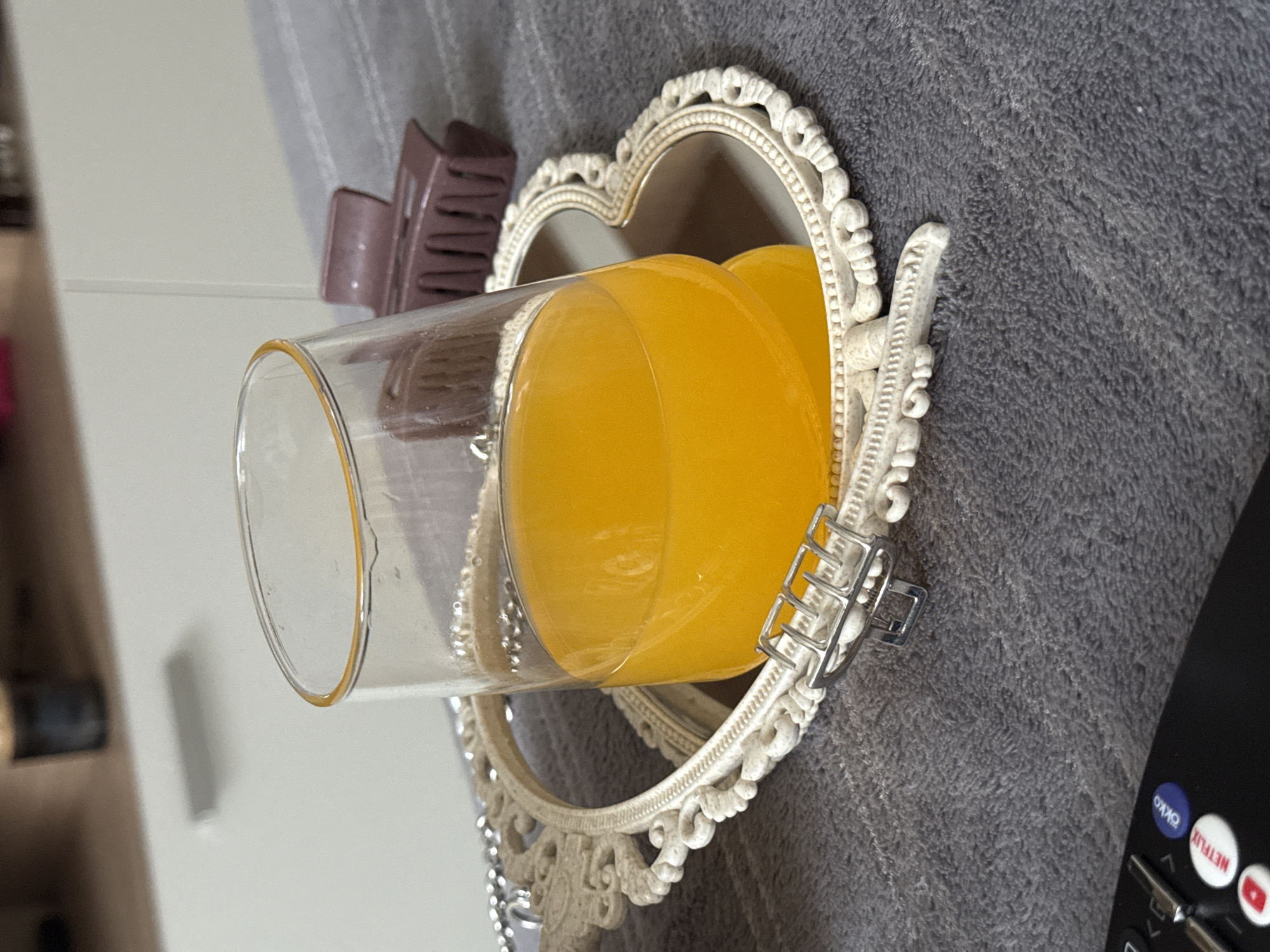 A glass of orange juice is resting inside a heart-shaped ornate mirror. A brown hair clip is situated next to the glass on top of the mirror. The mirror rests on a gray, textured surface, possibly a blanket or rug. A black remote control with various buttons is visible on the lower right side of the frame. The scene appears indoors, possibly on a bed or sofa.