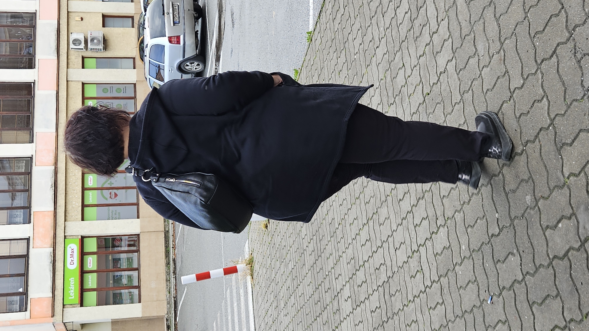 A person, viewed from behind, stands on a paved walkway in front of a building with the sign "Lekáreň Dr.Max" in Prešov, Slovakia. The person is wearing a dark hooded jacket, dark pants, and dark shoes. A black handbag is slung over the person's shoulder. A silver station wagon is parked to the right of the building. A red and white striped pole is visible on the left side of the walkway. The building has multiple windows and air conditioning units on its facade. The walkway is made of interlocking grey pavers.