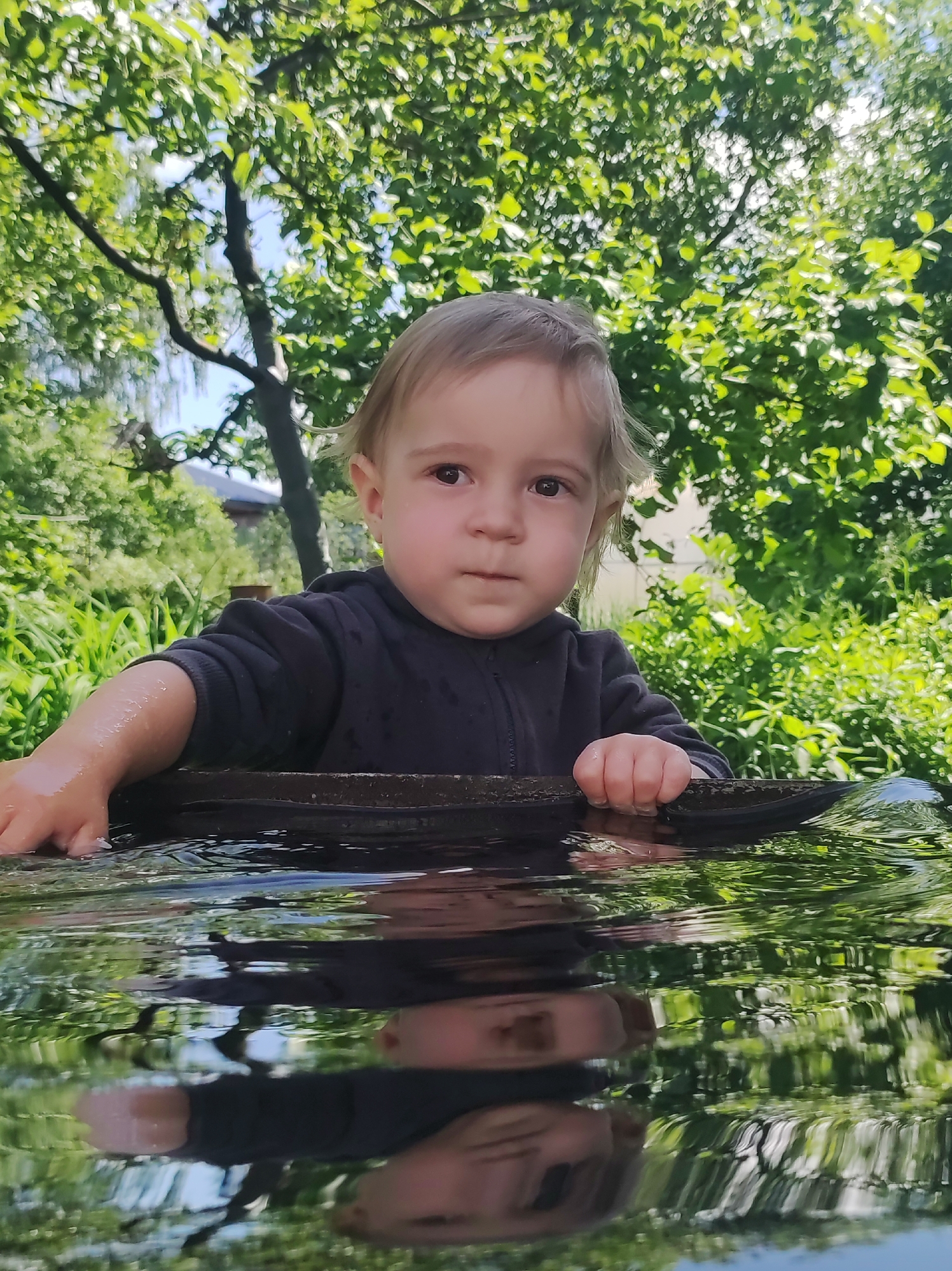 This vibrant portrait features a young child, likely a toddler, with light-colored hair and dark eyes, immersed in an outdoor natural setting. The child, wearing a dark, long-sleeved garment, is positioned at what appears to be the edge of a water body or container. Their left arm is visibly wet with water droplets, and both hands are resting on a dark, rounded rim just above the water's surface, suggesting playful interaction with the water. The child's reflection, including their face and upper torso, is clearly visible in the calm water directly below the rim, creating an interesting visual dynamic.
The background is a lush tapestry of bright green foliage, consisting of trees and bushes, rendered with a soft blur that emphasizes the child. The natural light filtering through the leaves suggests a sunny daytime setting, casting a pleasant glow on the scene. The child's expression is one of calm curiosity or contemplation, looking directly ahead or slightly towards the viewer. This capture conveys a moment of childhood exploration and connection with nature in Chaikovsky, Russia. There is no visible text in the image.