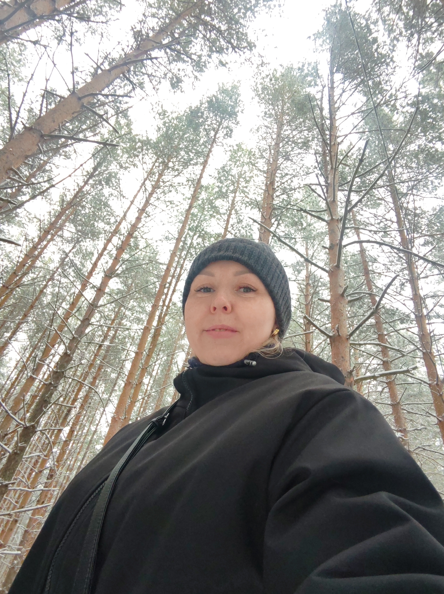 A low-angle, full shot depicts a woman in a snowy forest. The woman is centered, wearing a black beanie and a black jacket with a shoulder bag strap. The backdrop features numerous tall, slender trees stretching upwards, their branches covered in snow. The sky is overcast. The photo appears to have been taken in a forested area in Chaikovsky, Russia.
