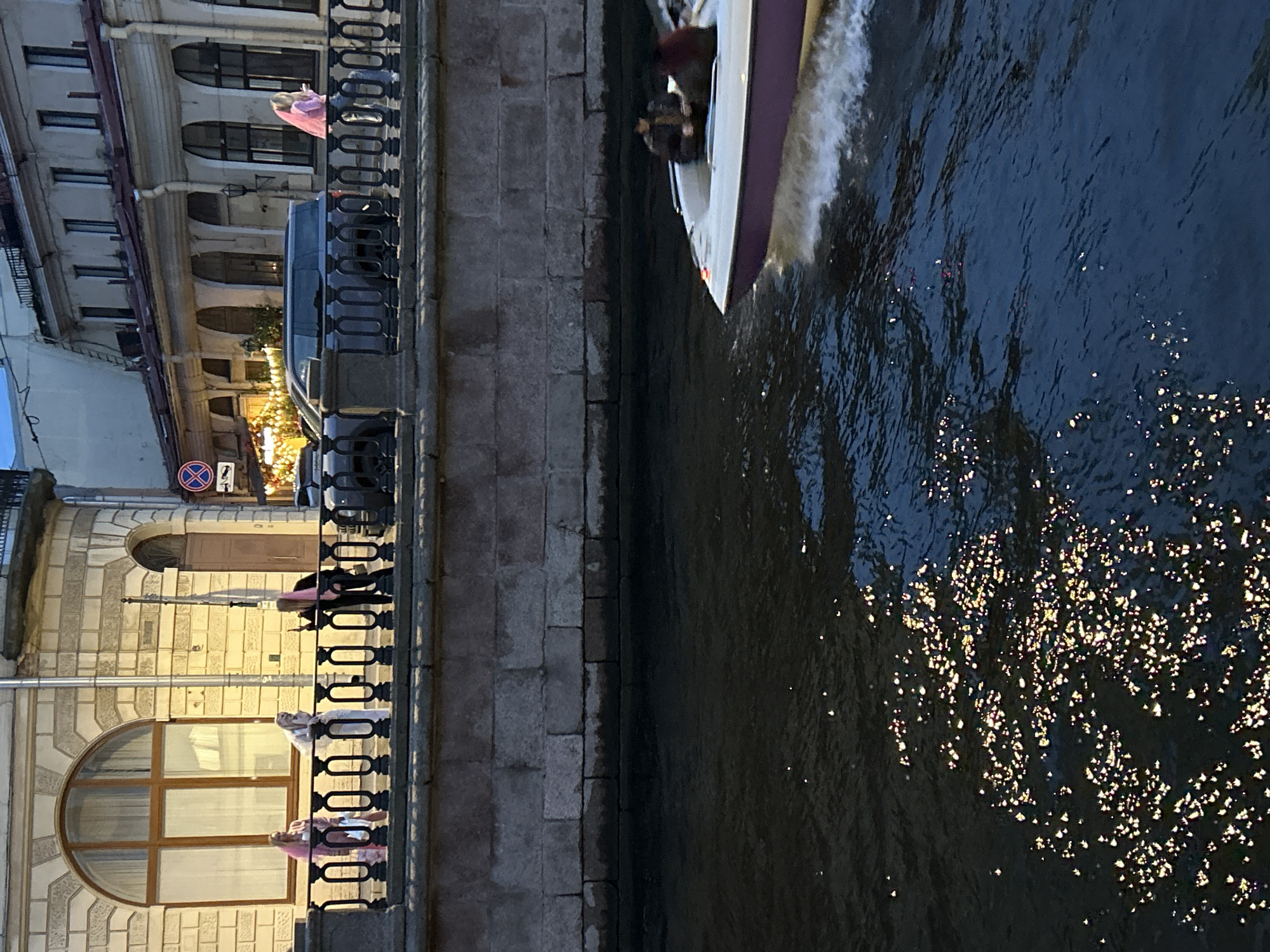 A wide-angle, vertical photograph captures a scene along a canal, likely in an urban setting with historical architecture. The primary subjects are a boat moving on the water and people on a balcony or walkway overlooking the canal.
In the foreground, on the left, a building with a detailed facade features arched windows and a decorative railing. Several people are visible on the walkway behind the railing. One person, wearing a pink garment, leans over the railing, possibly observing the activity on the water. Further down, two other individuals, one in white and another in pink, are also present. The architecture suggests an older European city.
To the right, the canal dominates the frame. A small white and purple motorboat is cutting through the dark water, creating a wake. The boat appears to be carrying passengers, with at least one person visible inside. The surface of the water is textured and reflects light, creating a sparkling effect, particularly in the lower right quadrant of the image. This suggests the time of day might be dusk or evening, with artificial lights reflecting off the water, or a sunny day with the sun at a low angle.
On the left side of the image, behind the building facade, hints of activity can be seen, including what appears to be illuminated festive lights and parked vehicles. A traffic sign with a red circle and a horizontal line is also visible.
The overall mood is tranquil, with the dynamic movement of the boat contrasting with the static architecture and the reflective calm of the water. The lighting suggests it is either late evening or early morning, and the weather appears to be calm.