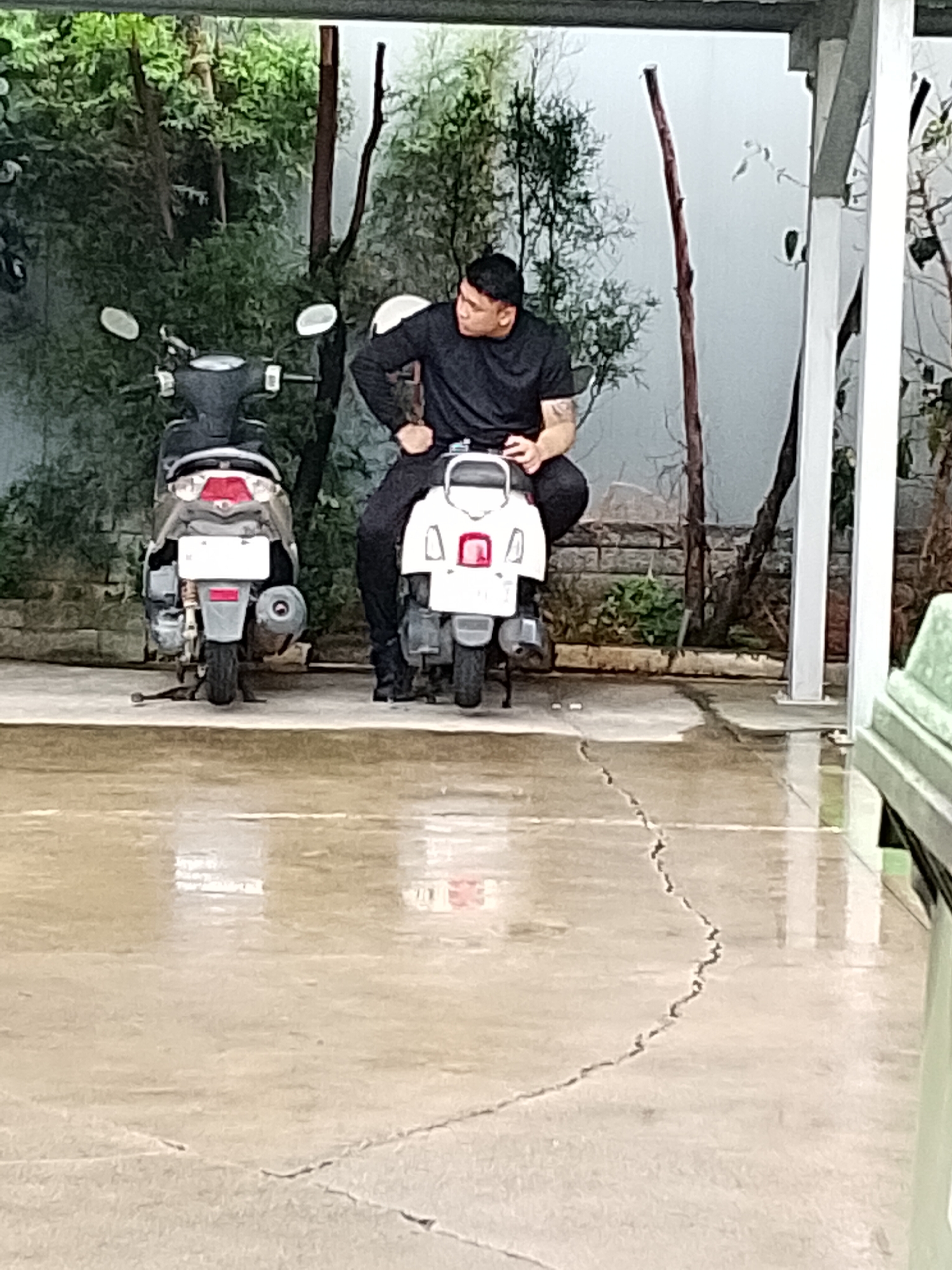 A man is sitting on a white scooter. He is wearing a black t-shirt and black pants. He is looking to the right. To his left is a dark colored scooter. The scooters are parked on a wet concrete surface. In the background, there are trees and a brick wall. A white pillar is visible on the right side of the image. The scene appears to be an outdoor area, possibly a parking lot or a covered walkway.