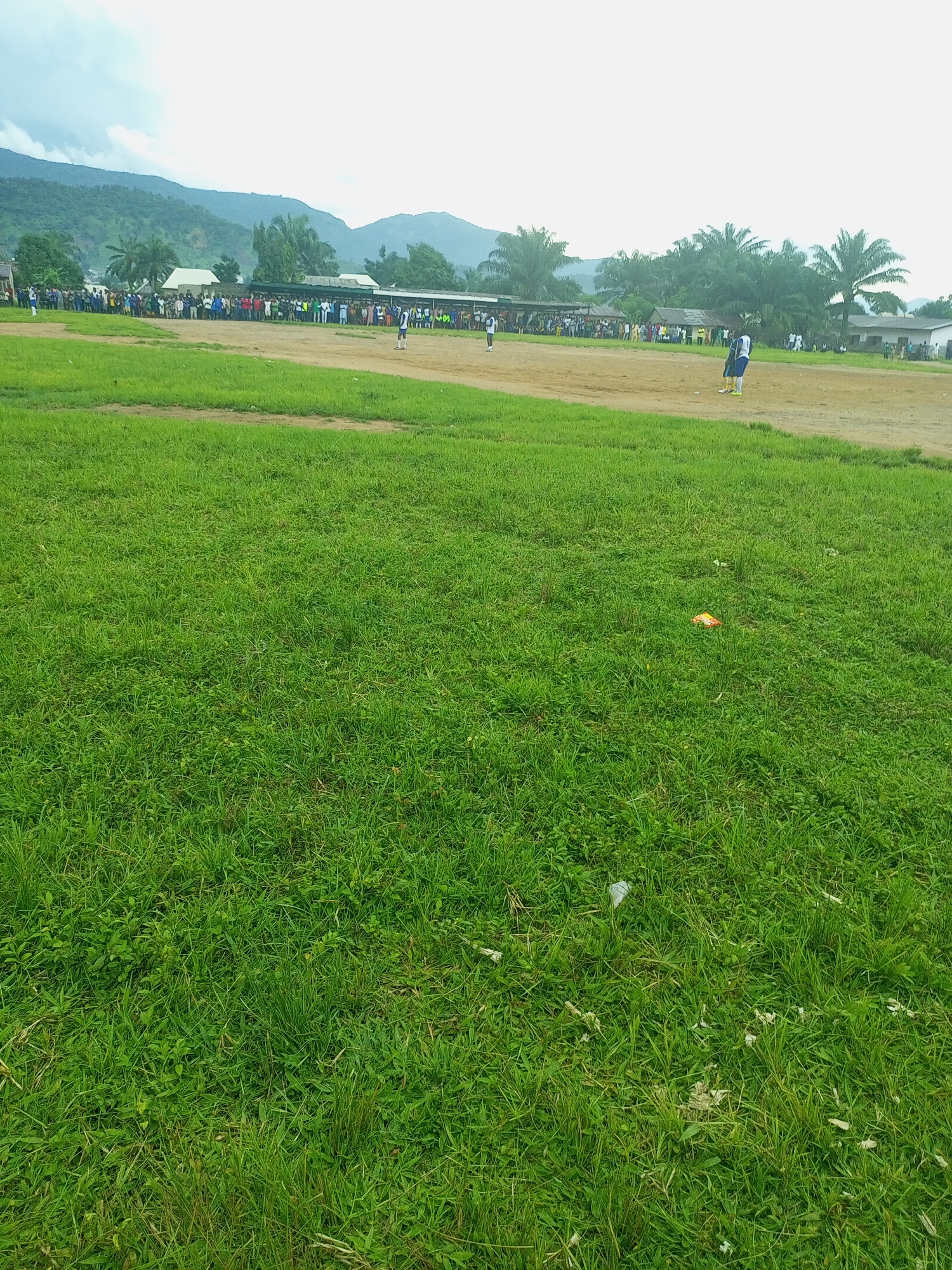 The image depicts a wide, grassy field in Serti, Nigeria, likely a public area or a school ground, given the presence of a crowd gathered in the background along a dirt path. A group of people are standing and watching an event unfolding on a dusty, brown section of the field. Two individuals, possibly athletes, are visible on this section of the field, one in a blue and white uniform and another in a white uniform. In the background, there are buildings and lush green vegetation, including palm trees, with a range of mountains forming a backdrop against a hazy sky. Scattered on the grass in the foreground are a few pieces of litter, including a crumpled orange wrapper and some white plastic. The overall scene suggests an outdoor event or gathering taking place in a community setting in Nigeria.