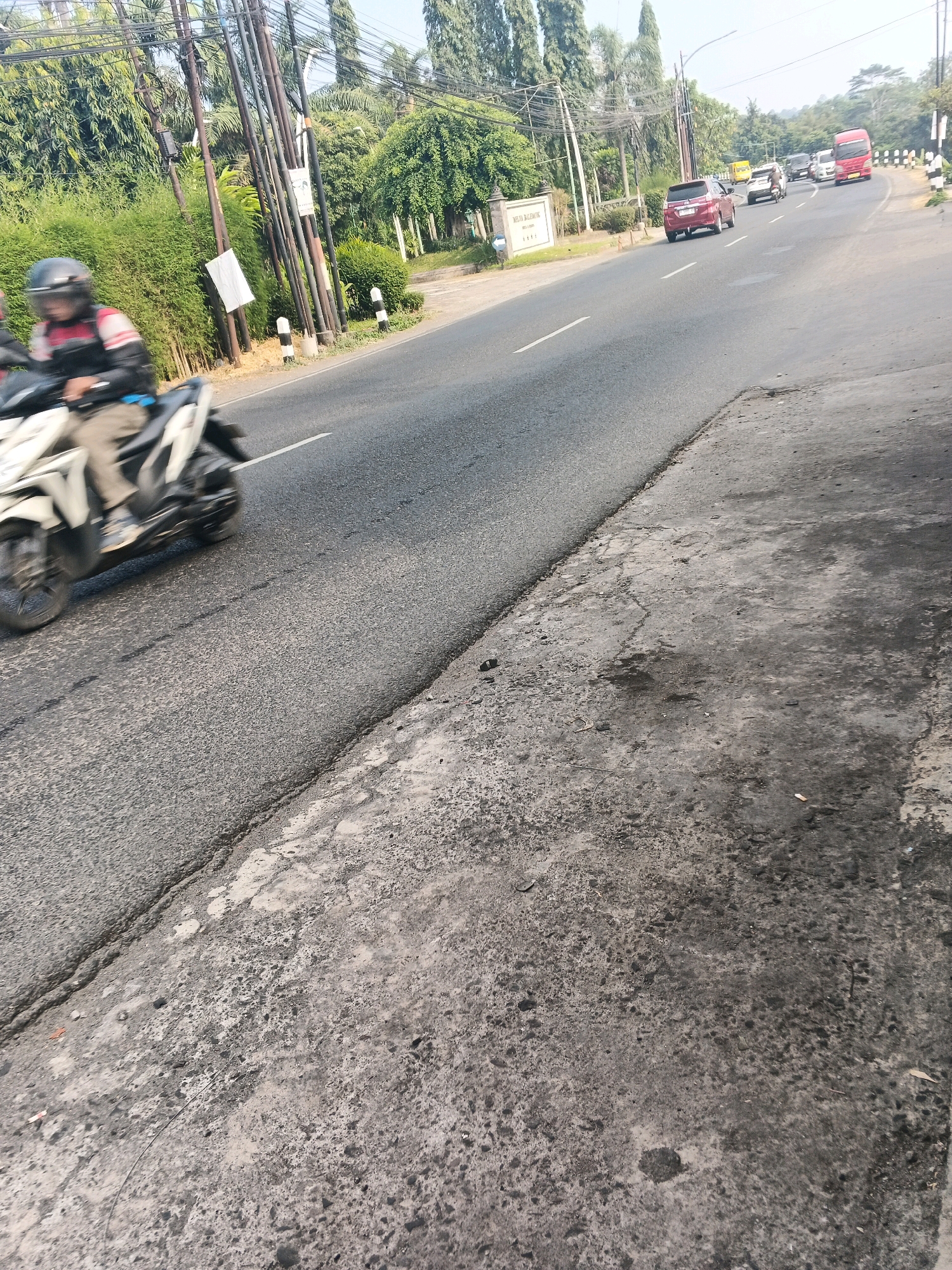 The image shows a street scene in Ungaran, Indonesia. A motorcyclist in a helmet and jacket is moving down the road, blurred due to motion. Several cars and a red bus are also visible on the asphalt road. The road is bordered by a concrete sidewalk with some debris on it. Lush green trees and utility poles line the roadside. A sign with Indonesian text is also present on the left side of the road. The sky is clear, suggesting it is daytime.