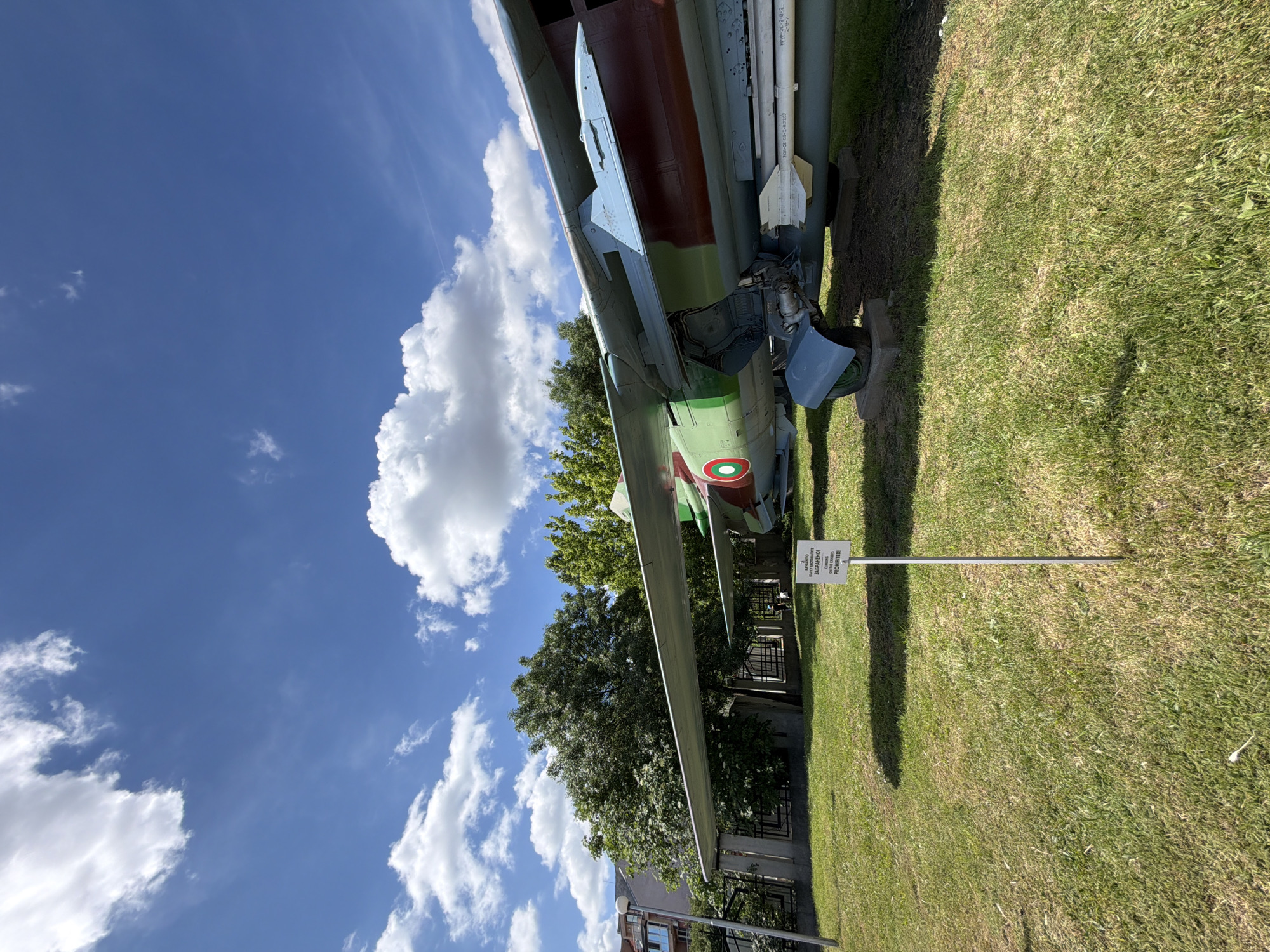 A static display of a military aircraft, likely a Soviet-era jet fighter, is presented outdoors under a bright blue sky with scattered white clouds. The aircraft, painted in a camouflage scheme of green, brown, and blue, is positioned on a grassy lawn. Part of a wing extends across the frame, revealing a missile mounted beneath it. In the foreground, a white sign on a metal pole displays text in Bulgarian and English, reading: "КАРАНДЕТО ИГРАТА И ДРУГИ ВЪНШНИ ИГРИ СА ЗАБРАНЕНИ! WARNING: PLAYING AND OTHER OUTDOOR GAMES ARE PROHIBITED!". The aircraft appears to be part of an outdoor exhibition, possibly at a museum, in Sofia, Bulgaria. Behind the aircraft, trees and a building are visible. The scene is well-lit by sunlight, casting shadows on the grass. There are no people visible in the image.