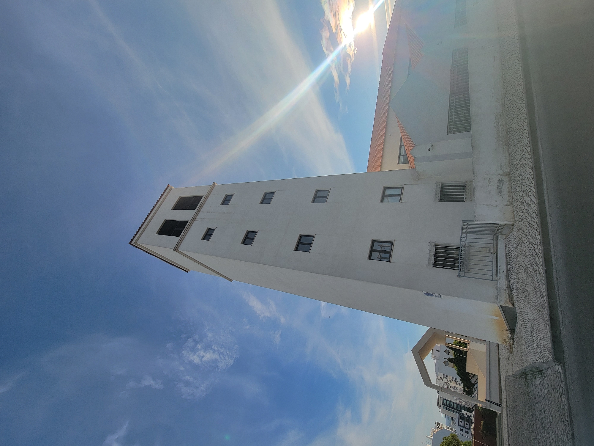 The image shows a modern, white, multi-story tower with dark-framed windows, located in Quarteira, Portugal. The tower has a distinct, angled design. Adjacent to the tower is a building with a red-tiled roof. In the background, several other modern buildings are visible, along with trees. The sky is blue with scattered white clouds, and the sun is shining brightly, creating a lens flare effect across the upper right portion of the image. The ground in front of the buildings is paved with cobblestones and asphalt. A metal fence is present in front of some windows on the lower level of the tower. The overall scene depicts an urban environment under a clear sky during daylight.