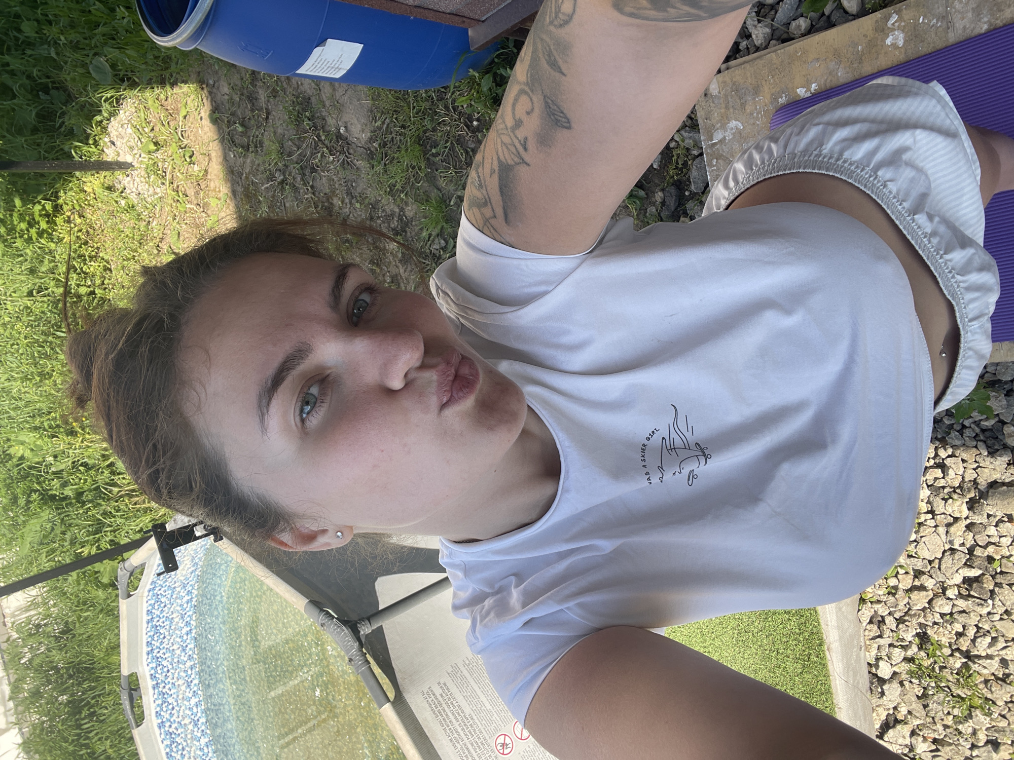 A woman takes a selfie outdoors, captured from an overhead angle looking down. She has brown hair pulled back, green eyes, and is making a playful "kissing" face. She wears a white t-shirt featuring a black graphic illustration of a person skiing and the text "NAS A SKIER GIRI" (likely "HAS A SKIER GIRL"). Tattoos are visible on her left arm, and she has a navel piercing. She is also wearing light-colored, possibly striped, shorts.
The setting appears to be a sunny backyard or garden area, characterized by green grass and patches of gravel or small rocks. In the background, a blue barrel, possibly for water storage or part of a portable pool system, is visible. Text on the barrel includes safety warnings in English and French regarding "POOLS," "ACTIVE ADULT SUPERVISION," "FENCING REQUIREMENTS," and "PISCINES PORTABLES." To the left, part of an outdoor chair or lounger with a patterned fabric (blue and white dots) and some readable warning labels is present. A purple mat, possibly a yoga mat, is visible beneath her lower body on the right. The overall impression is casual and relaxed. The location context is Мосметаллоконструкция Онт, Russia.