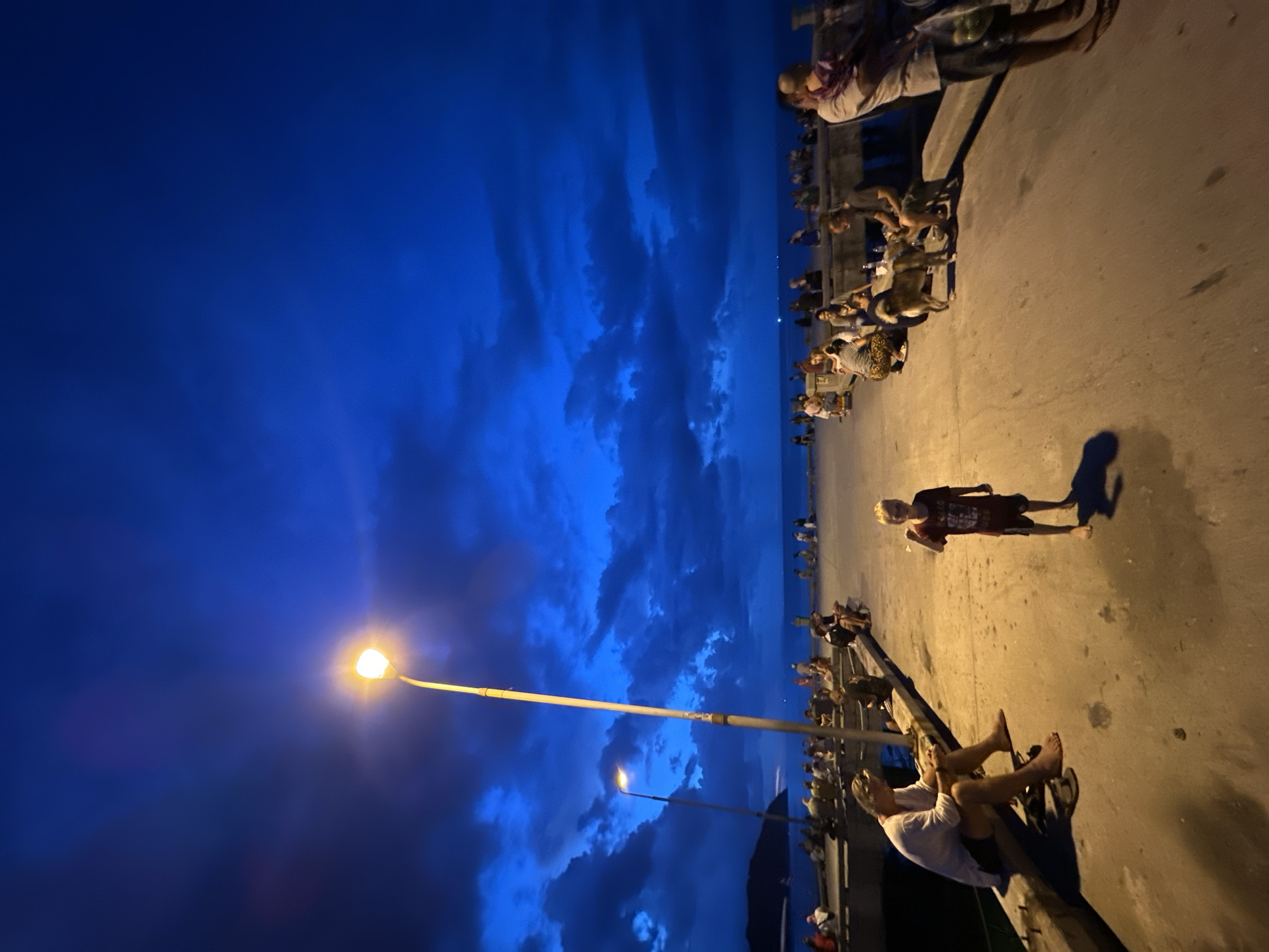The image captures a scene at dusk on a pier in Koh Phangan, Thailand. A boy stands in the foreground, facing the camera with his right fist raised. The pier is populated with numerous people sitting and standing, many facing the ocean. A lamppost illuminates the area, casting long shadows. In the background, the ocean is visible under a deep blue, cloudy sky. Several boats are spotted on the water in the distance. The overall atmosphere is relaxed, with people enjoying the evening by the sea.