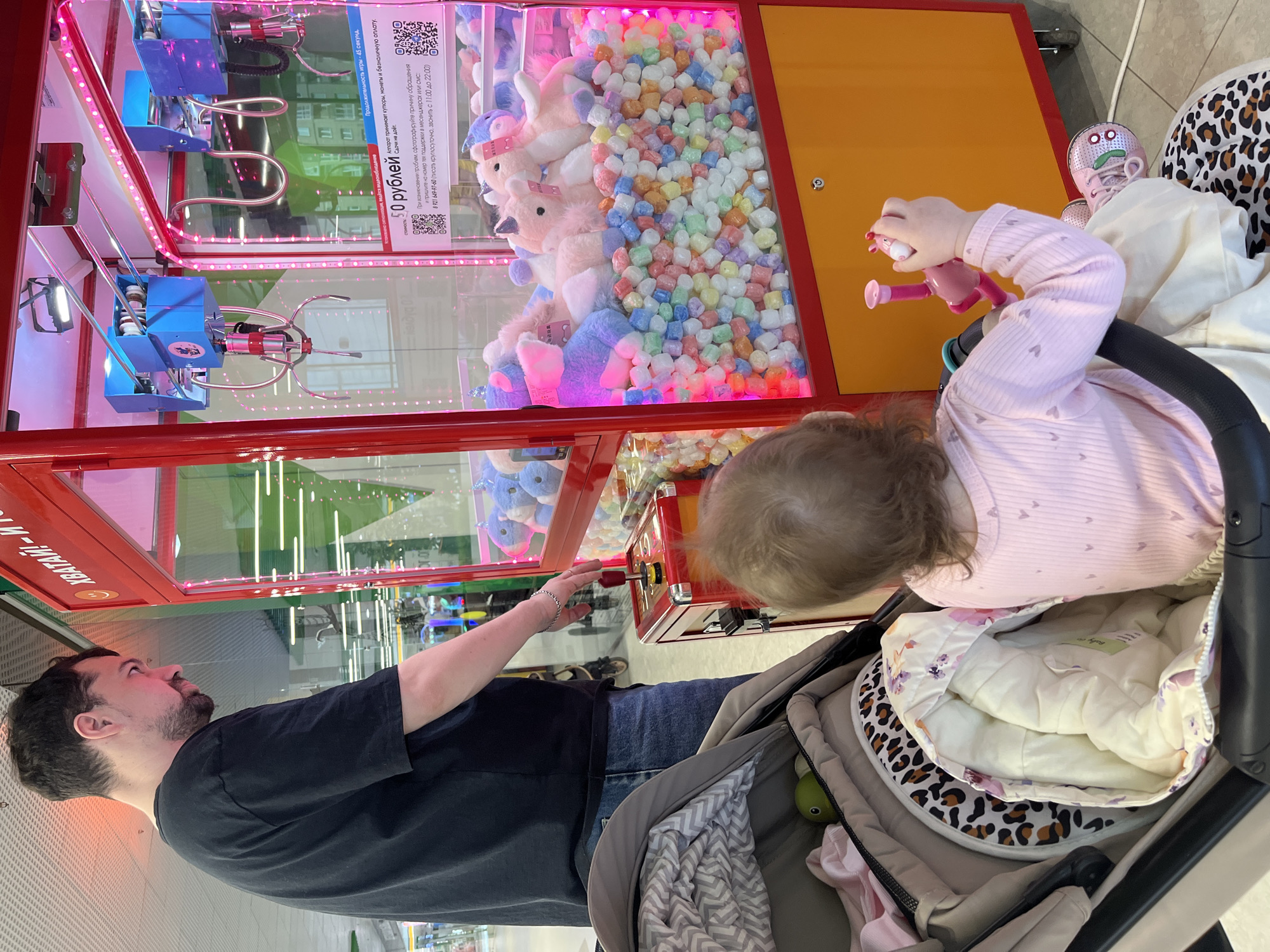 A medium, eye-level shot shows a man and a child in front of a claw machine, probably located in Shushary, Russia based on the text in Russian on the machine. The man, with a dark beard and wearing a black t-shirt and jeans, is looking at the machine and operating the joystick. The child, wearing a pink long-sleeved shirt, is sitting in a stroller. The stroller also contains a blanket and other items. The claw machine is red with a glass front, filled with plush toys and colorful balls. The machine displays the price of 50 rubles and instructions in Russian. The background shows an interior shopping mall with lights and stores visible.