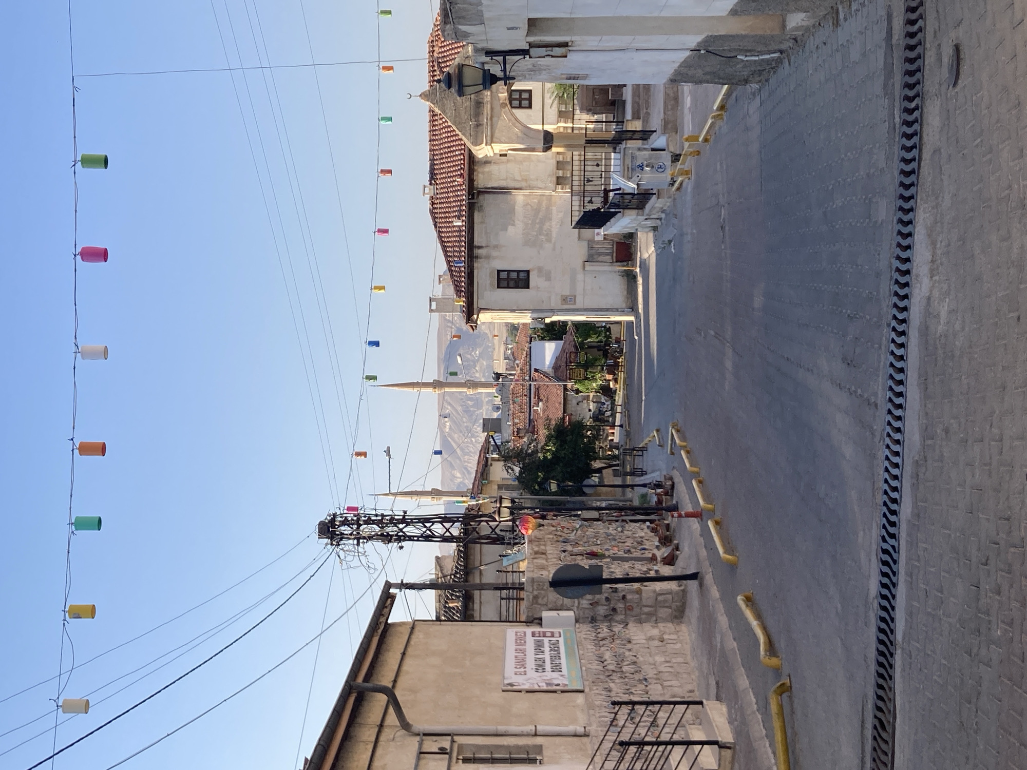 The image depicts a street scene in Avanos İlçe Merkezi, Turkey, characterized by traditional architecture and overhead decorative elements. The street itself is paved with interlocking stones in the foreground, transitioning to asphalt in the middle ground. Yellow, curved bollards line the edges of the asphalted section. A drainage grate runs across the width of the street in the lower portion of the image.
Buildings on either side of the street exhibit a consistent architectural style, featuring stone facades, tiled roofs, and visible windows. On the left, a building has a stone wall adorned with what appear to be small decorative items, possibly pottery, and a sign that reads "EL SANATLARI MERKEZİ ÇOMLEK YAPIMI DENEYEBILIRSINIZ" (Handicrafts Center, You Can Experience Pottery Making). A metal railing and steps lead up to this building's entrance.
On the right, buildings also feature stone and stucco elements with tiled roofs. A traditional lamppost is mounted on a wall. In the background, the silhouette of minarets from mosques can be seen against the clear blue sky, suggesting a religious or historical context for the area. Further in the distance, rolling hills are visible.
Suspended overhead by wires are numerous colorful, cylindrical lanterns in various shades of yellow, green, orange, and pink, adding a festive or decorative touch to the streetscape. Electrical wires and a utility pole are also prominent features in the upper portion of the image. There are no people visible in the scene. The overall atmosphere is one of a quiet, sunlit day in a town with a distinct cultural heritage.