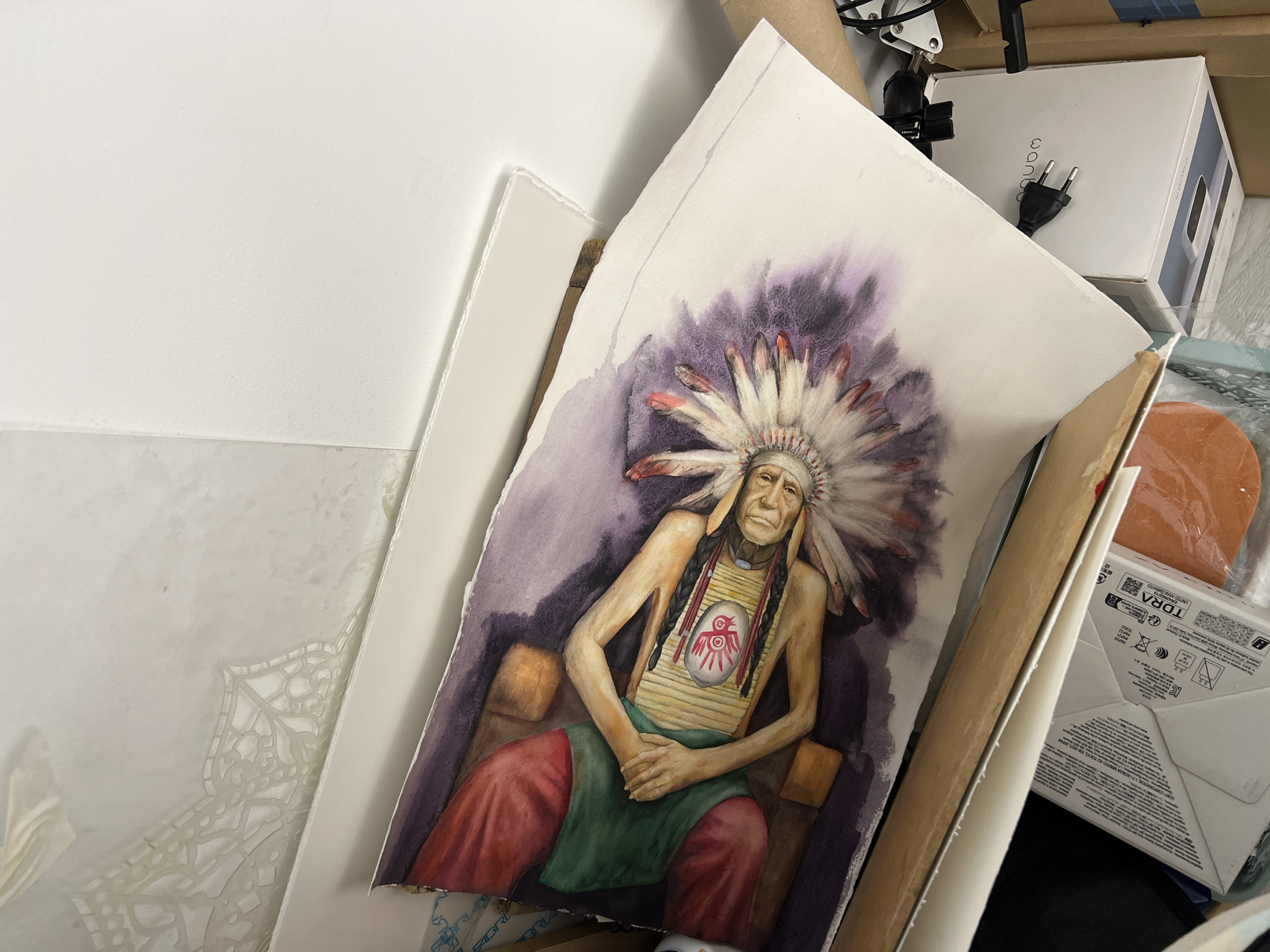The image shows a watercolor painting of a person wearing a Native American headdress, lying down with their hands resting on their chest. The person is depicted with braided hair and a smiling expression. In the center of their chest, there is a circular emblem with a red handprint and a bird-like symbol. The background of the painting is a blend of purple and white watercolor washes. The painting is placed on a surface that appears to be a table or a shelf, with other objects visible in the background, including cardboard boxes and what looks like electrical plugs. The lighting in the scene is bright, suggesting an indoor environment. The specific location within the city of Unknown, Unknown cannot be determined from the provided image.