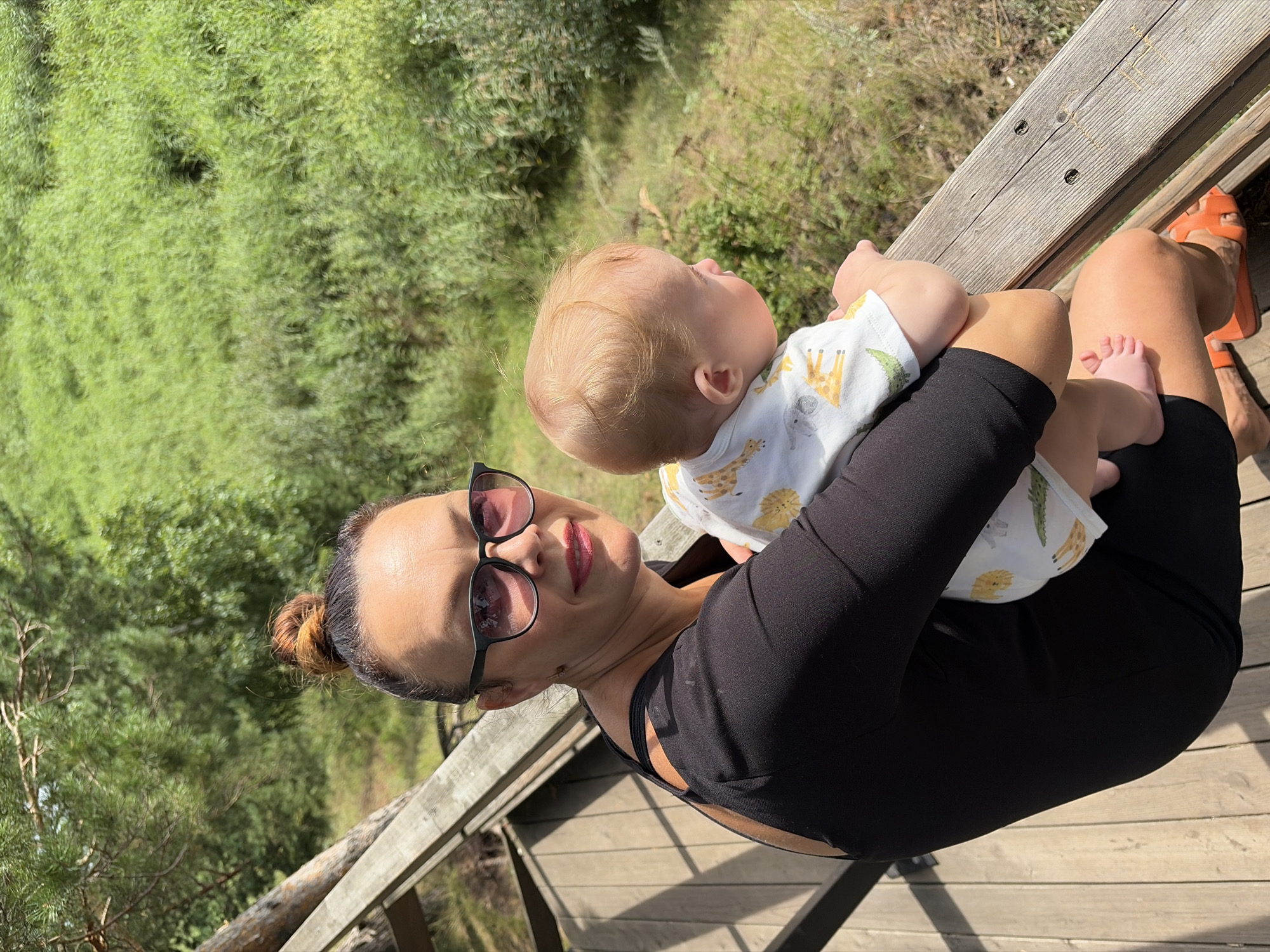 A woman with her hair in a bun and wearing sunglasses holds a baby while sitting on a wooden deck. The woman is wearing a black dress and orange sandals. The baby is wearing a white onesie with a safari print. They are outdoors with a background of lush green foliage. The image appears to be taken on a sunny day. The location is likely Votkinsk, Russia, given the context of the request.