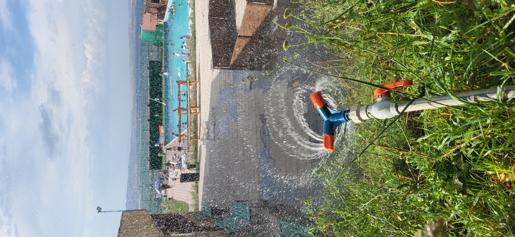 In Сырымбет, Kazakhstan, an outdoor scene unfolds under a partially cloudy sky. In the foreground, a garden hose nozzle, a blue Y-shaped sprinkler with orange tips, is actively spraying water in a circular pattern onto a paved area. The spray creates a mist that catches the sunlight. Lush green grass and some flowering plants surround the nozzle.
Beyond the immediate foreground, a paved walkway leads towards a swimming pool area. Several people are visible, some relaxing by the pool and others in the water. There are lounge chairs and umbrellas set up. A dark trash receptacle is situated near the walkway. To the left, a building with a textured, light brown facade is partially visible. In the background, a fence encloses the pool area, and beyond that, rolling hills covered in green vegetation extend towards the horizon. The overall impression is a recreational area on a sunny day, with the sprinkler suggesting a method of cooling or watering the surroundings.