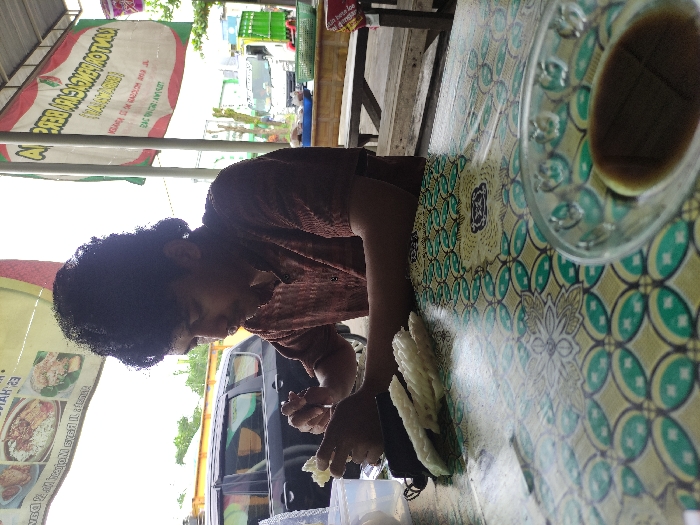 This media file captures a casual, outdoor dining scene in Kepanjen, Indonesia, during daytime. The image is oriented sideways (rotated 90 degrees counter-clockwise).
The primary subject is a person with dark, curly hair, wearing a dark brown patterned shirt, seated at a table. They are leaning forward, looking down, and appear focused on an activity involving their hands, which are holding or interacting with several white, elongated items resembling crackers (likely krupuk).
The table is covered with a distinctive tablecloth featuring a green, yellow, and white batik-style pattern. On the right side of the table, a clear glass cup holds a dark beverage, possibly coffee or tea. A white plastic container or bowl is also visible near the person's hands.
In the background, a large white banner with red and green accents is prominently displayed, suggesting it's an advertisement for the eatery. Visible text on the banner includes "KOTOKAN GEBREK," which appears to be a name or type of dish, along with phrases like "KOLOSAL OLEH RASANYA" and other smaller, less legible text. The banner also features images of various food dishes. Behind the person and the banner, a dark-colored vehicle, possibly a minivan or SUV, is partially visible, indicating a roadside or open-air restaurant setting. The lighting suggests it is daytime, possibly overcast or brightly lit. The overall scene conveys a casual dining experience.