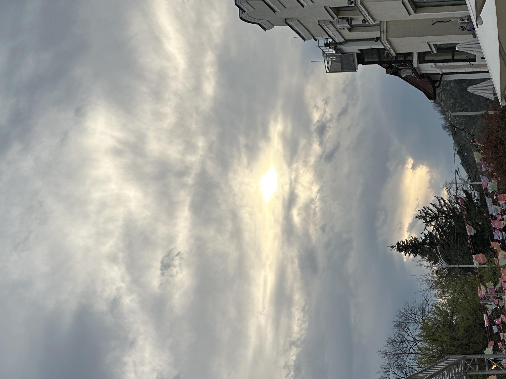 A vertical, upward-angled shot captures a partially obscured sun amidst a heavily clouded sky, adjacent to sections of architecture and foliage, likely taken in or near the city of Plovdiv, Bulgaria. A light-colored building is visible at the upper right of the frame. Green trees and some other vegetation are visible, including decorative pennants strung from poles, suggesting an event. Parts of a scaffolding structure are visible at the bottom.