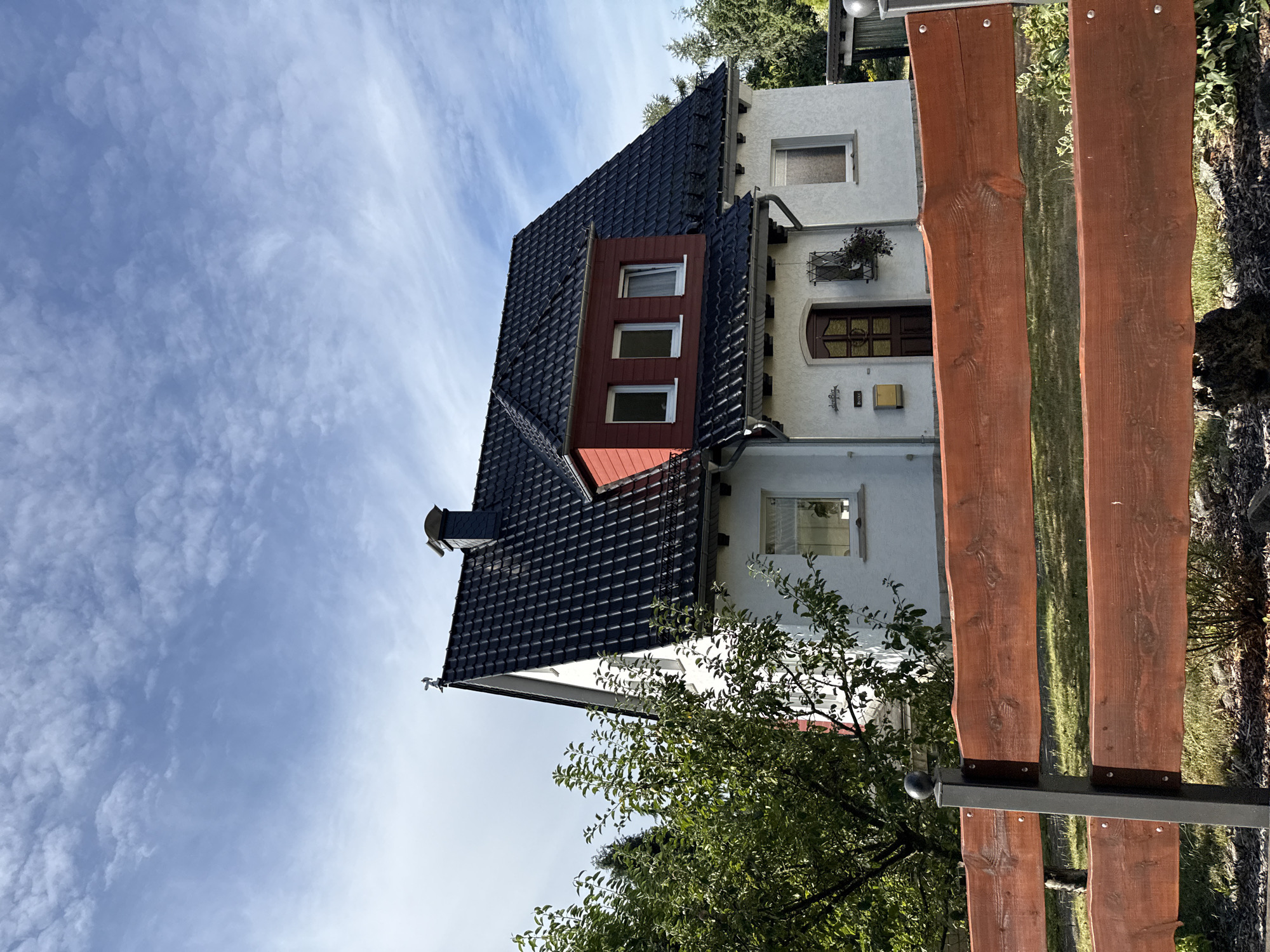 This is an exterior, eye-level, full shot of a two-story house with a dark tiled roof, white exterior walls, and a red dormer window. The house is situated in a suburban or rural area. In the foreground, there is a wooden fence with dark metal posts. Behind the house, there are trees and a clear blue sky with scattered white clouds. The house has a dark brown front door, a mailbox, and several windows. A small weather vane is visible on the peak of the roof. The overall impression is of a well-maintained residential property on a clear day. The location is identified as Bad Sachsa, Germany.