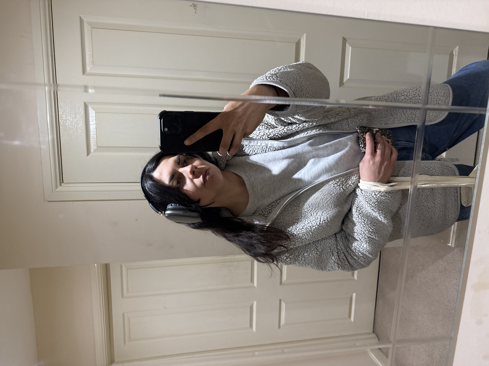 A woman wearing headphones takes a mirror selfie. She is indoors, likely in a bathroom, based on the cabinet reflected in the lower right of the mirror. She wears a light grey, fleecy jacket over a light grey t-shirt. She holds a black smartphone in her right hand and a small wallet or card holder in her left. She is wearing blue jeans. The background features white paneled doors. The specific location within the city of Great Yarmouth is not discernible from this image.