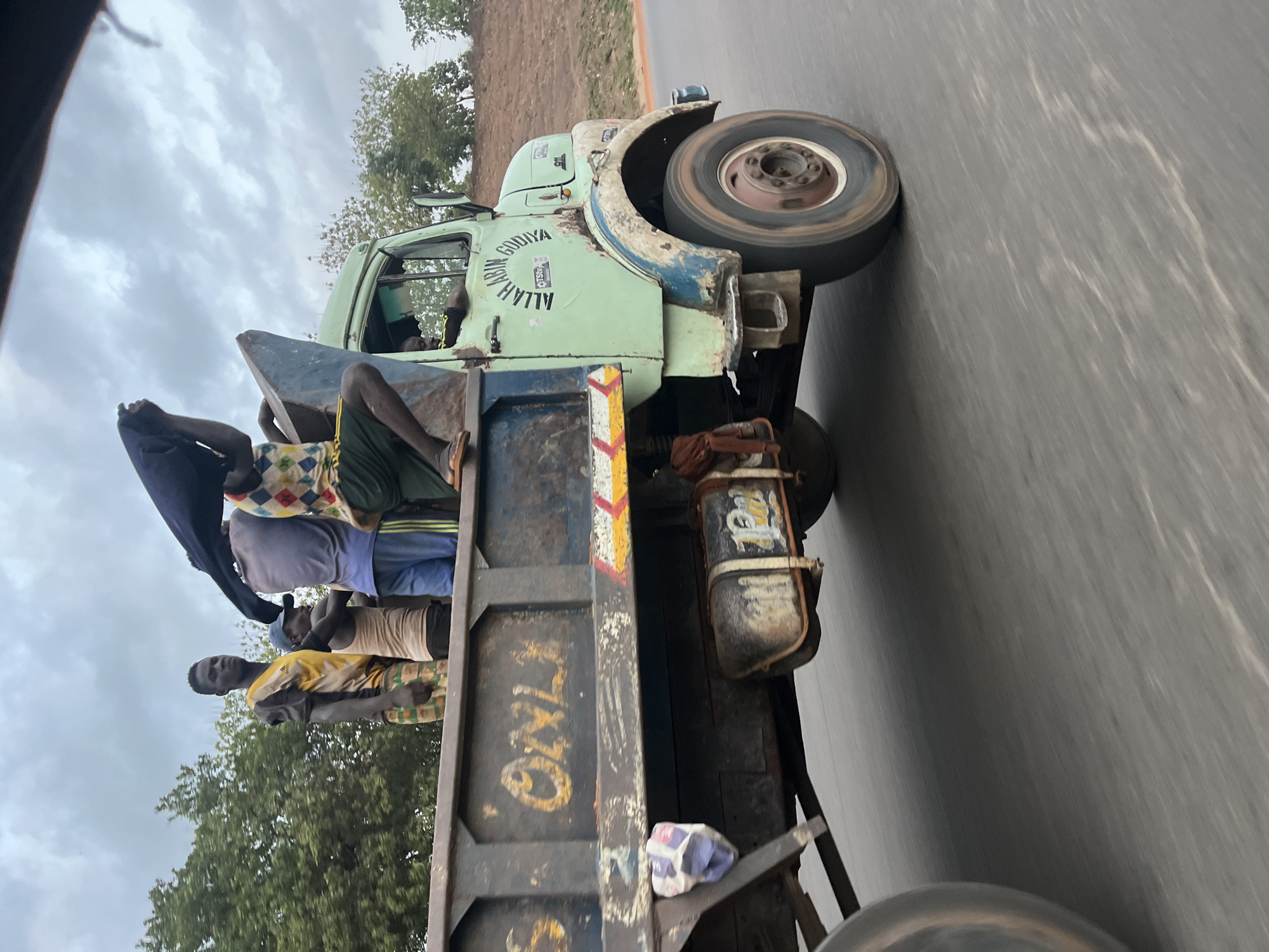 The image depicts an old, light-green truck moving on an asphalt road. The truck is filled with multiple people, who appear to be riding in the back. The truck has visible wear and tear, including rust and damage to the body. Text, including "ALLAH", is visible on the side of the vehicle. The environment suggests a location in Kuchingoro, Nigeria, with trees and vegetation in the background. The sky is overcast.