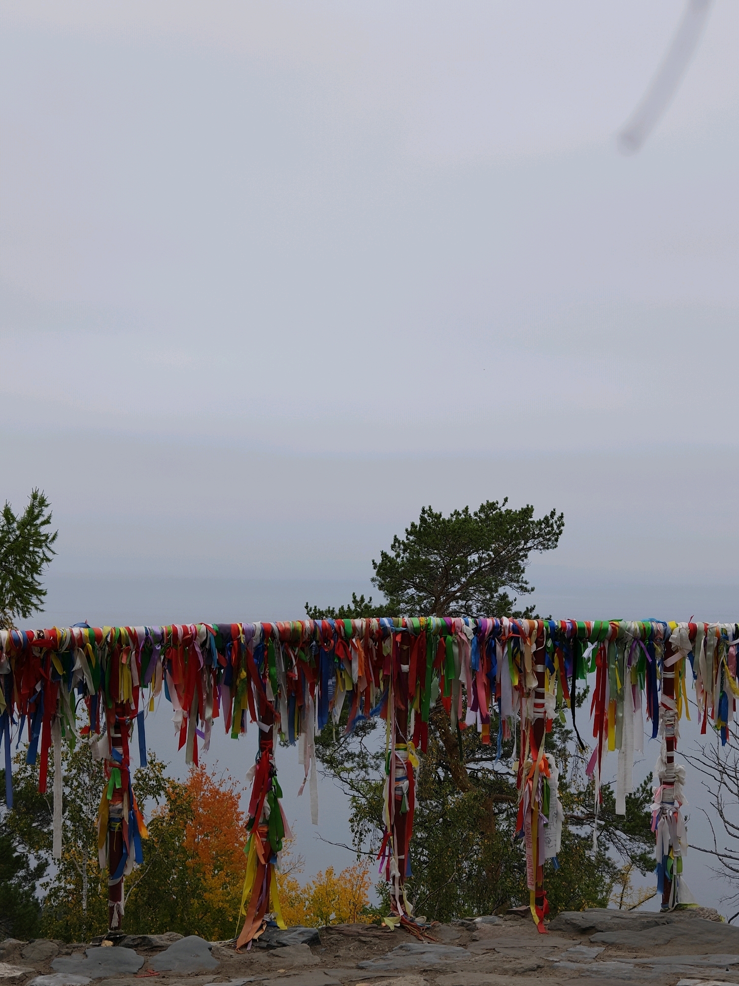 The image shows a horizontal pole decorated with numerous colorful ribbons tied to it. These ribbons are of various colors, including red, blue, yellow, green, white, and purple. They are of different lengths and appear to be made of fabric. The pole is supported by two vertical posts, which are also adorned with ribbons. Behind the pole, there is a landscape with trees, some of which have autumn foliage in yellow and orange colors. The sky is overcast and gray. In the distance, there appears to be a body of water, possibly a lake or sea, and a hazy horizon. The scene is set outdoors, on a rocky surface in the foreground. The presence of prayer ribbons tied to a pole is a common practice in spiritual sites, particularly in Buddhist and Shamanistic traditions. The text visible on some of the ribbons, though not fully legible, suggests a spiritual or commemorative purpose. While the exact location within Nikola, Russia, is not specified, the imagery is consistent with natural or sacred sites often found in such regions.
