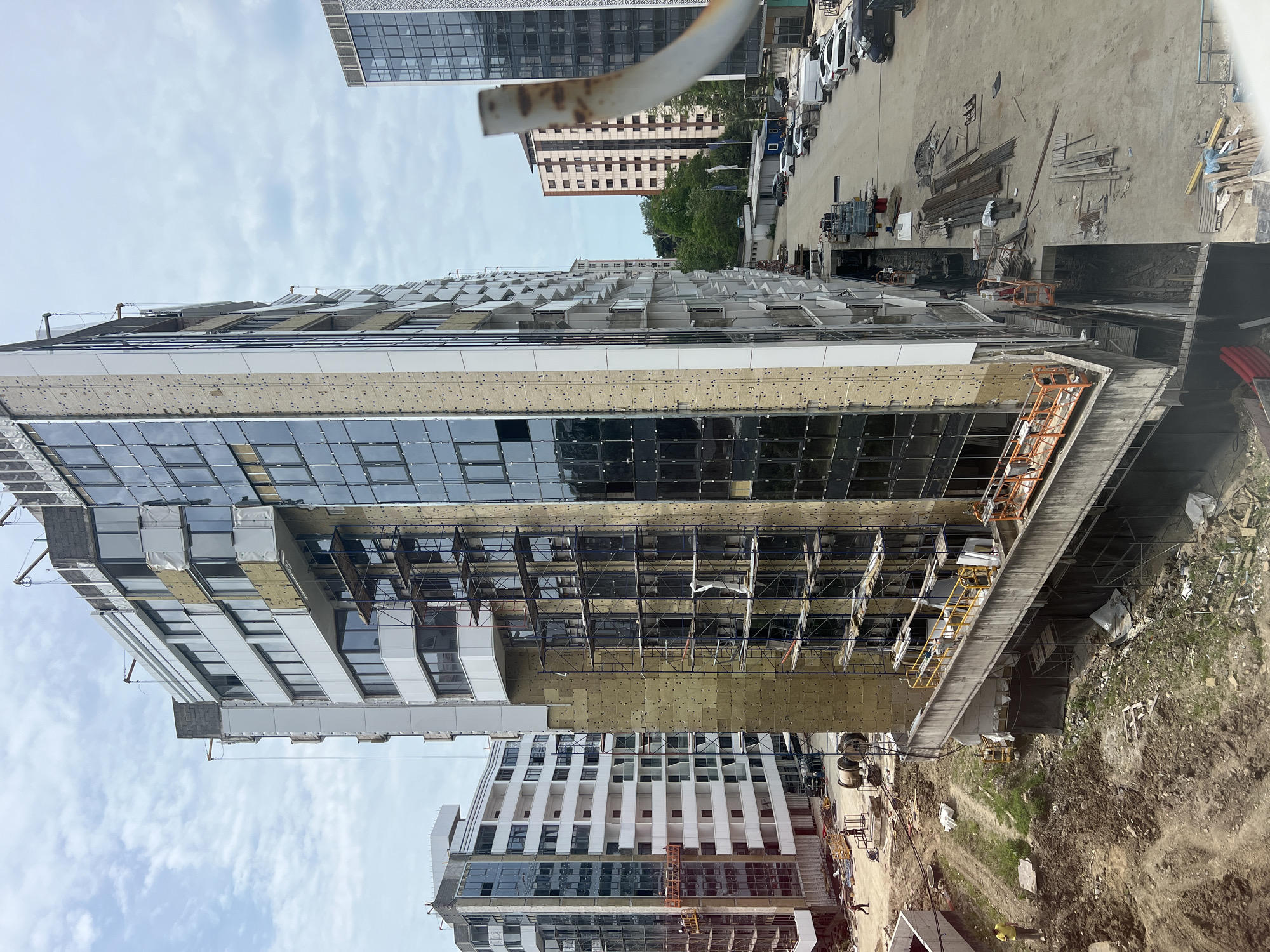 The image depicts a construction site in Sochi, Russia, characterized by several multi-story buildings in various stages of development. The foreground is dominated by a large building undergoing exterior work, with scaffolding attached to its facade. This building features extensive glass windows and exposed insulation material. To the left, another building, appearing to be residential, is nearly complete with white balconies and dark windows. The ground level is rough and unpaved, with piles of dirt, construction debris, and stacks of building materials such as rebar and lumber. Several vehicles are visible in the background, parked along a paved road that runs alongside the construction area. The sky is overcast with scattered clouds. Two individuals are visible on the construction site, one walking on the dirt ground and another on a platform attached to the scaffolding of the main building.