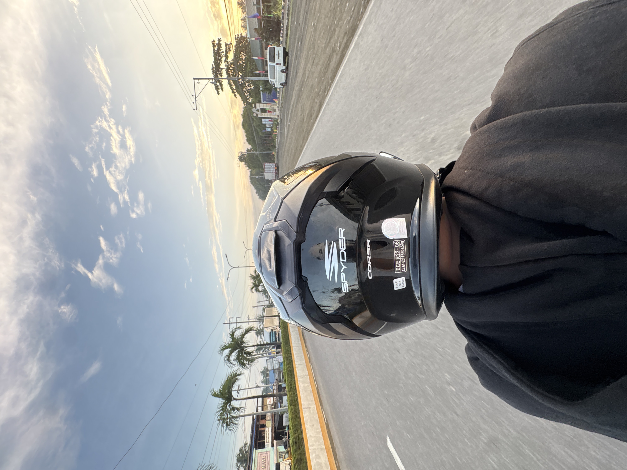 The image is a first-person perspective taken from the back of a motorcycle, likely in Carmen, Philippines, during a sunset. The rider is wearing a black motorcycle helmet with the brand "SPYDER" visible, and a black hooded jacket. The helmet also has a sticker that reads "ECE R22-06 XL 61-62 1550±50g" and "FINAL QC PASSED".
The road ahead is paved, with a concrete barrier on the left side of the road. Along the left side of the road, there are buildings with businesses and a few palm trees. On the right side of the road, there's more vegetation, including trees and some flags. The sky is a light blue with scattered white clouds, illuminated by the warm tones of the setting sun. Utility poles and wires are visible in the distance.
Traffic is present, with a white van and other vehicles on the road. The image captures a moment of travel on a public road during a scenic time of day.