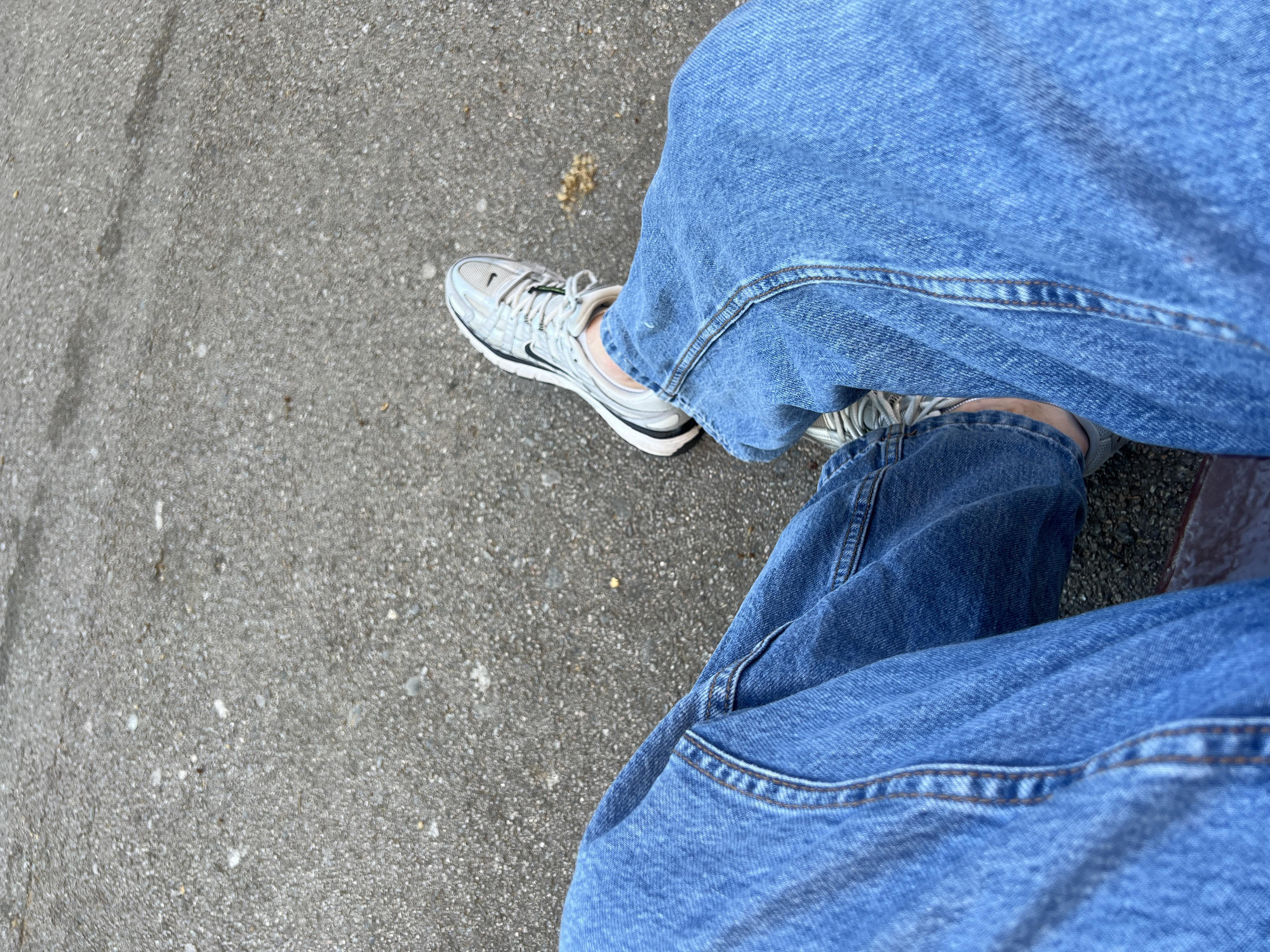 This media file captures a high-angle view of a person's lower legs and feet, likely from a first-person perspective. The individual is wearing blue denim jeans, characterized by visible seams and a slightly relaxed fit. Their feet are shod in light grey or silver athletic sneakers featuring black accents, a white sole, and a prominent black Nike swoosh logo on the side of the left shoe. The shoelaces are white with distinct green aglets.
The setting appears to be outdoors, with the legs positioned over a grey, textured pavement, consistent with asphalt or concrete, which has a slightly speckled appearance. A small cluster of dried, light-brown plant material is visible on the ground near the left foot. Below the right leg, a dark, horizontal edge, possibly a bench or a step, is partially visible, suggesting the person is seated or resting.
The lighting is bright and even, indicating daytime conditions, possibly under an overcast sky or during a period of diffused sunlight, as there are no harsh shadows. The image does not contain any explicit visual cues to identify the location as Barcelona, Spain, beyond the provided context. The overall impression is casual and relaxed. The only clearly discernible text is the Nike swoosh logo on the sneaker.