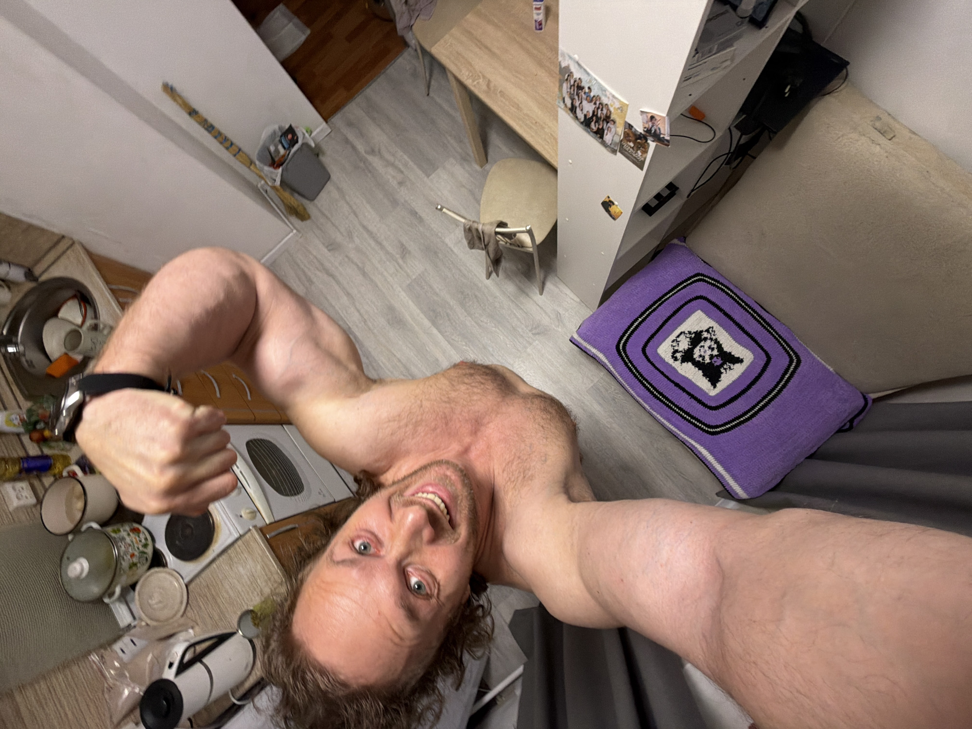 The image is a high-angle selfie taken by a shirtless man with light brown, curly hair and blue eyes, smiling broadly at the camera. He displays a muscular physique and wears a black watch on his left wrist. The setting is an indoor residential space, likely a small apartment or studio in Saint Petersburg, Russia, given the provided context.
The scene combines kitchen, dining, and living areas. In the foreground, a kitchen corner features a white electric stove, various pots and mugs, a white kettle, and a partially visible sink with dishes. Adjacent to this, a light wooden dining table with a chair, over which a piece of clothing is draped, is positioned. Next to the table is a white shelving unit or cabinet adorned with several small photographs. Further into the background, a light-colored sofa is visible, featuring a distinctive purple crocheted pillow with a white and black geometric pattern. Gray curtains are drawn on the right side of the frame. The floor throughout the space is covered in light grey wood laminate. Other household items include a long, stick-like object (possibly a broom) leaning against a white wall, next to a small grey trash can. The man's cheerful expression suggests a confident and playful mood. There is no discernible text visible in the image. The lighting appears to be artificial, making the time of day indeterminate.