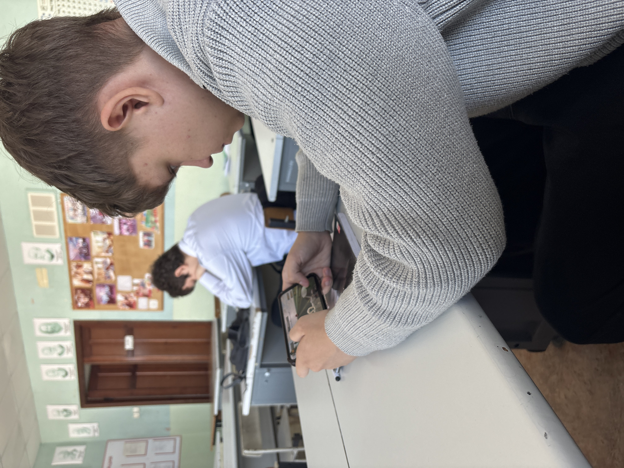 The image captures an indoor scene, likely a classroom or study environment, featuring two young males. The perspective is angled, showing the subjects and surroundings sideways.
In the immediate foreground, a young male with short brown hair is seen in profile, his gaze directed downwards. He is wearing a light grey, ribbed knit sweater and is holding a black smartphone horizontally with both hands. The phone's screen displays blurry content, possibly an outdoor scene with greenery. A white desk surface is prominent in front of him, exhibiting minor scuffs. His black lower garment is partially visible beneath his arm.
Further into the background, another young male is visible from behind, wearing a white long-sleeved shirt. He has dark, curly hair and is leaning over a light-colored desk, seemingly engaged in an activity on its surface.
The room features light green walls adorned with various items. A cork bulletin board is prominently displayed, covered with numerous small photographs or papers. Below the bulletin board, a wooden door is partially visible. To the left of the door, an empty wooden bookshelf with multiple shelves is mounted on the wall. Several white rectangular papers, possibly containing green drawings or text, are taped to the wall, along with another white board divided into smaller rectangular sections. The lighting suggests an indoor setting, but the time of day and weather are not discernible. No legible text is visible on any of the displayed papers or objects.