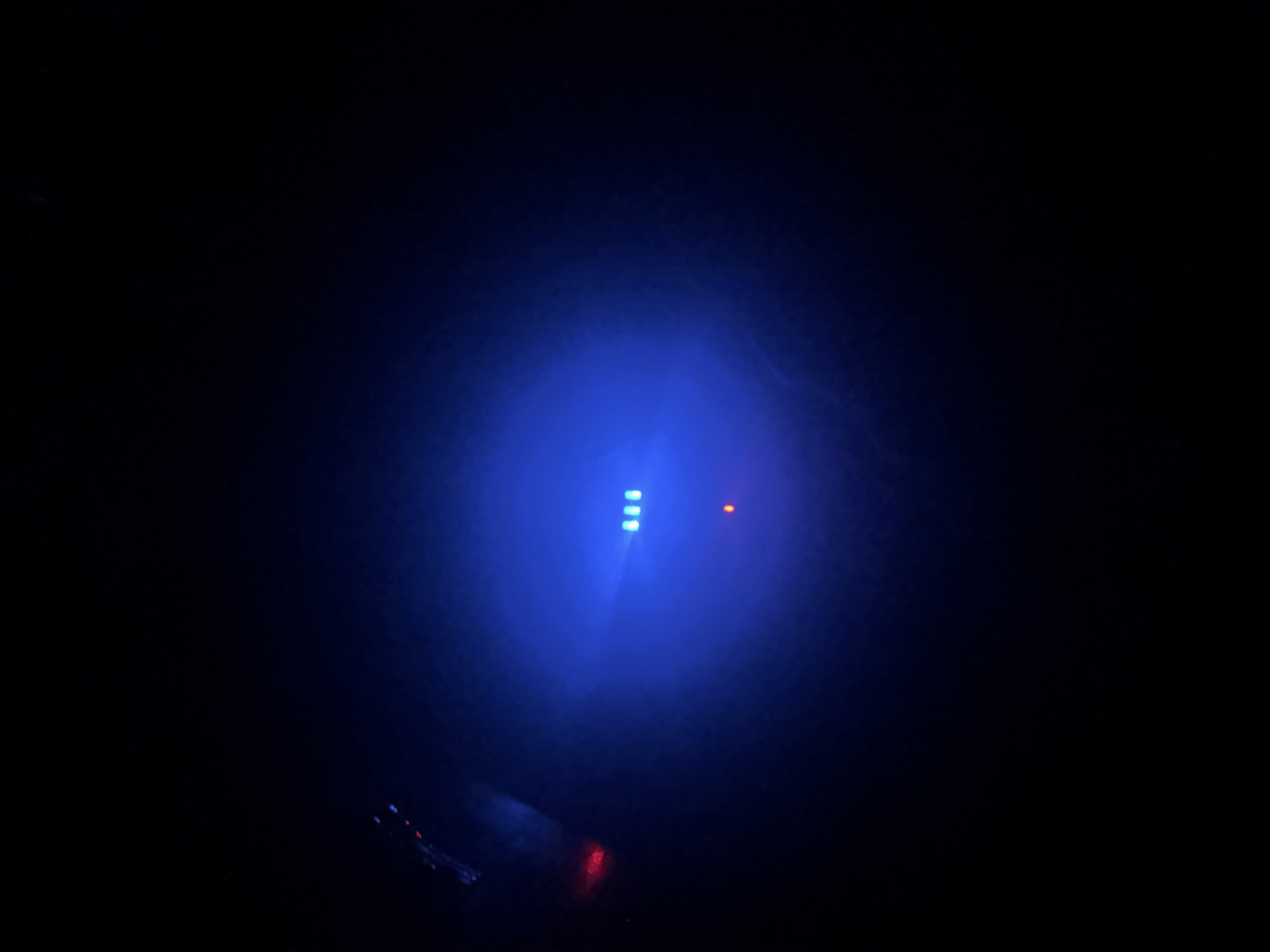 This media file is a dimly lit photograph captured in Ihiagwa, Nigeria. The primary subject is a series of small, glowing lights against a dark background. Three blue LED lights are arranged vertically in the center, with a single red light positioned to their right. There is also a faint red glow visible towards the bottom of the frame, and some blurred blue and white lights in the lower left corner. The overall scene suggests a low-light environment, possibly at night, with the lights potentially indicating the status of electronic devices or a control panel. The lack of other discernible subjects or environmental details makes it difficult to determine a specific activity or event, but the focus on the lights suggests they are significant. No text is visible in the image.
