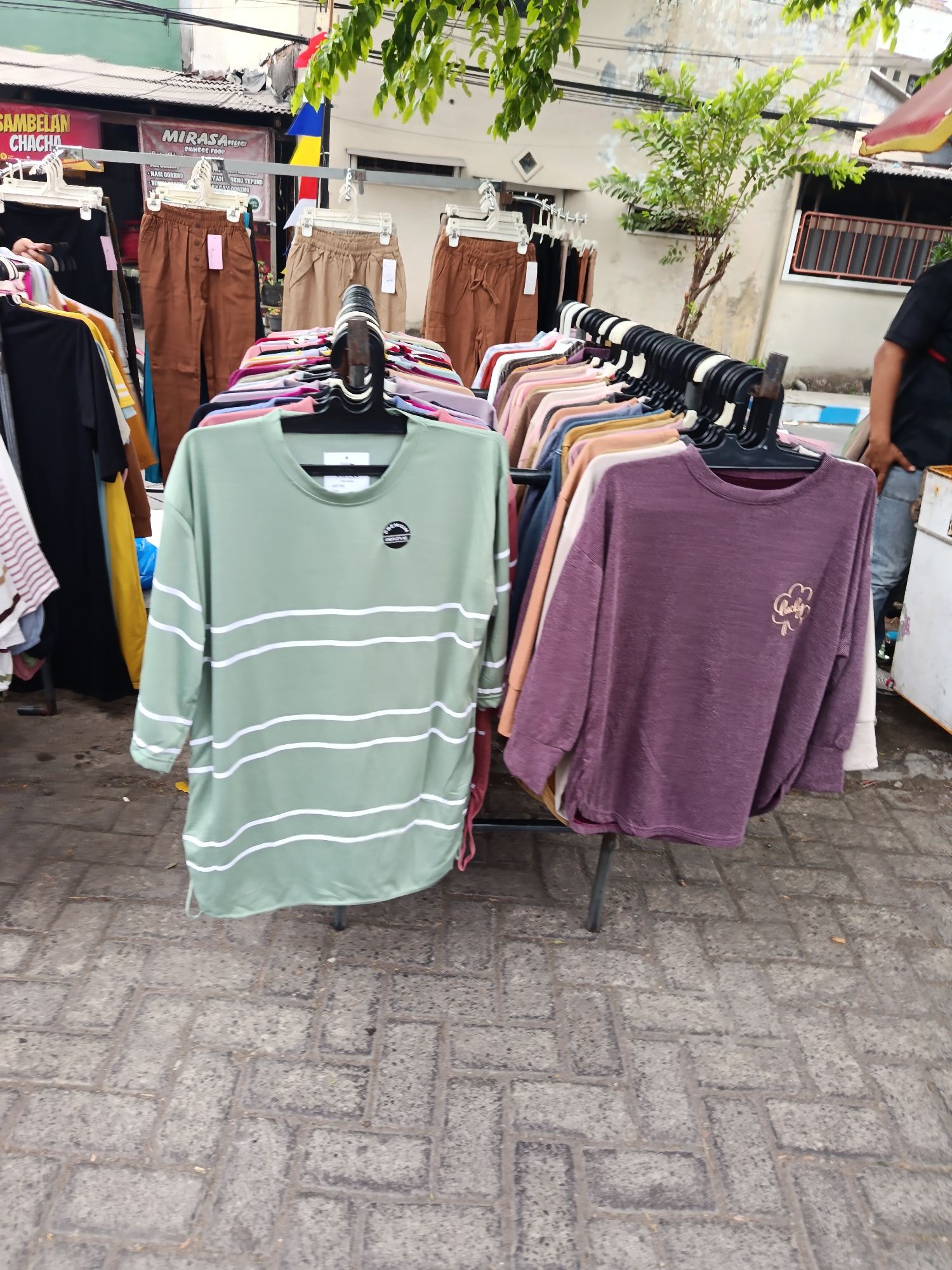 This is a full, eye-level shot of a clothing stall outdoors, possibly on a sidewalk, with a street visible in the background. The stall displays various garments, primarily t-shirts and pants, on hangers and racks. A light green t-shirt with horizontal white stripes is prominently featured in the foreground, alongside a purple t-shirt with a clover-like design. Behind these, a rack of folded shirts in various pastel colors is visible. Further back, brown pants hang on a rack, and a person's arm and shoulder are visible to the right, wearing a black t-shirt and jeans. The ground is paved with gray interlocking bricks. The background shows a building with a window and a red trim, and trees with green leaves overhead, suggesting an urban or semi-urban environment in Keboansikep, Indonesia.