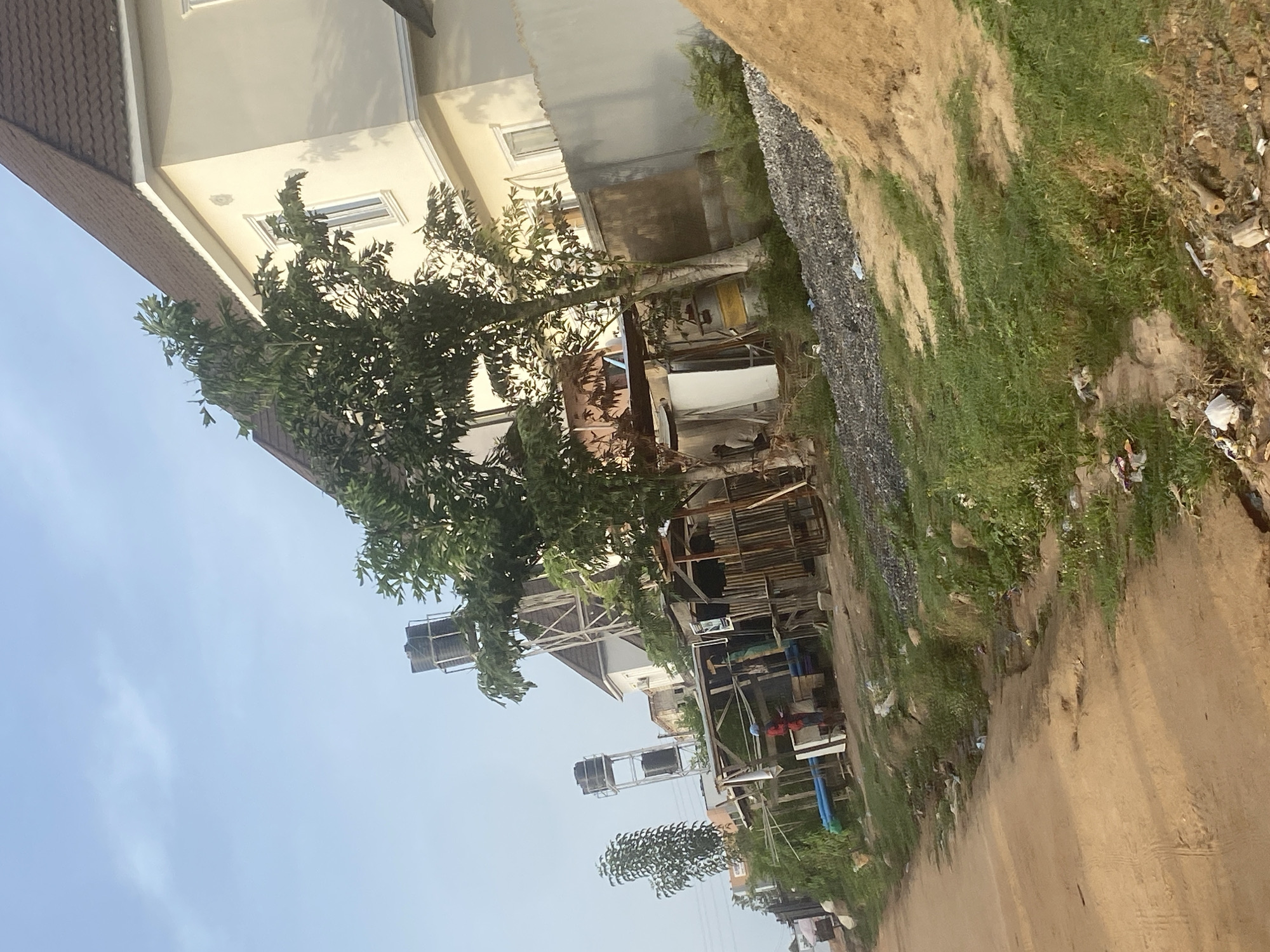 The image shows a street scene in Iddo Tudunwada, Nigeria. In the foreground, a sandy path or road is visible with patches of green grass and scattered debris along the edges. A modern, two-story building is visible on the left, constructed of light-colored materials and with a brown tiled roof. A large tree partially obscures the building. Further down the road, additional buildings and structures are visible, including what appears to be construction or storage areas with exposed frameworks. Overhead water tanks are supported by metal structures are also visible. The sky is light blue with some cloud cover.