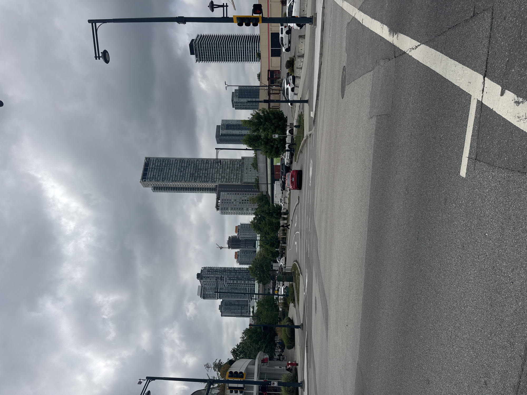 A street-level view of an urban intersection in Surrey, Canada, is captured under a cloudy sky. The foreground consists of asphalt road surfaces with visible lane markings, including a prominent white line diagonally traversing the lower right quadrant. In the middle ground, a street corner is visible with a curved building, a traffic light, a fire hydrant, and several trees. Beyond the street, a collection of high-rise buildings dominates the skyline, characteristic of a city center. Cars are present on the road and in parking areas. A lone pedestrian can be seen crossing the street in the distance. The overall atmosphere is overcast and urban.