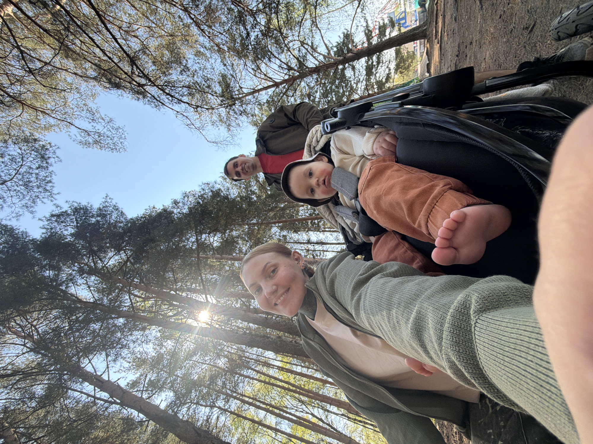 A family of three takes a selfie in what appears to be a stroller-filled area in Chaikovsky, Russia. The woman, holding the phone, is wearing a green jacket and a light-colored t-shirt. A man in a dark jacket and red shirt is positioned behind her. A baby in a stroller, dressed in beige and brown clothing, is prominently featured. Trees and blue sky are visible in the background, with dappled sunlight filtering through the branches. The foreground shows the stroller and a partial view of the woman's clothing.