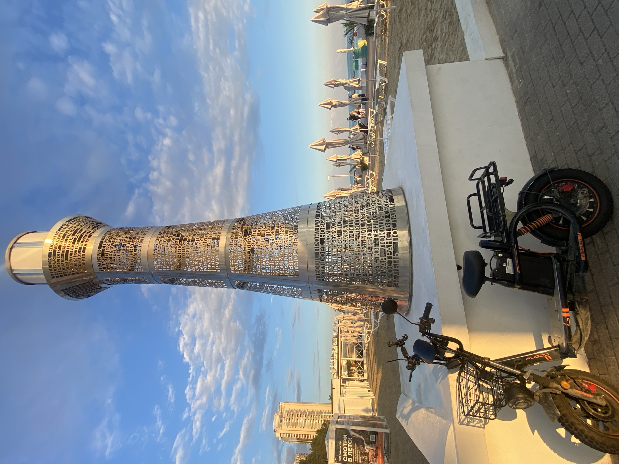 This is a vertical, outdoor photograph taken at what appears to be a beach resort in Sochi, Russia, during sunset. The golden hour light casts a warm glow across the scene.
The main subject on the left is a tall, cylindrical sculpture made of intricate metalwork with a repeating pattern of cut-out text. The text, visible in the provided crops, includes Russian words like "РОЗА ХУТОР" (Rosa Khutor), "СМОТРИ В ЛЕТО!" (Look in the summer!), and other phrases suggesting a resort or leisure destination.
In the foreground on the right, there are two electric scooters parked. Both are dark-colored with orange accents and have baskets in front. The scooter closer to the camera has "KUGO" visible on its frame.
The background shows a sandy beach with numerous closed beach umbrellas and lounge chairs, suggesting the day's activities are winding down. Beyond the beach, the sea is visible with a clear sky featuring wispy clouds. In the distance, a modern, high-rise building stands against the horizon.
The overall mood of the image is peaceful and serene, capturing the end of a day at the seaside. The presence of the sculpture and the scooters suggests a contemporary and amenity-rich location. The visible text confirms the location context as a Russian resort.