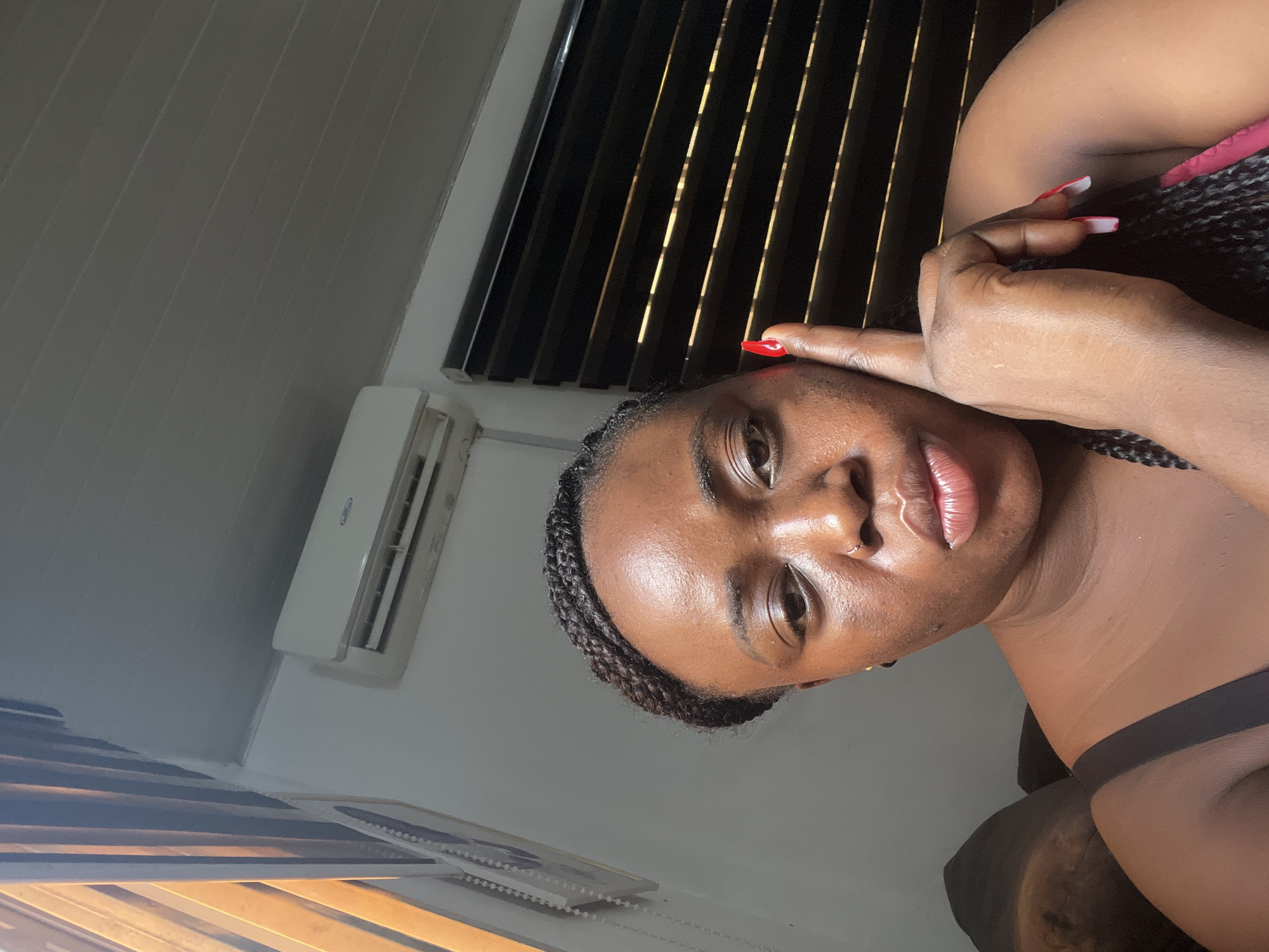 A close-up indoor shot of a Black woman with braided hair. She has a nose ring, reddish-pink lipstick, and red and white painted nails. She is wearing what appears to be a black top. Her right hand is near her face. In the background, there is a window with black blinds, an air conditioning unit, and part of a picture frame. The location is likely indoors in Abuja, Nigeria, although this is not explicitly shown in the image.