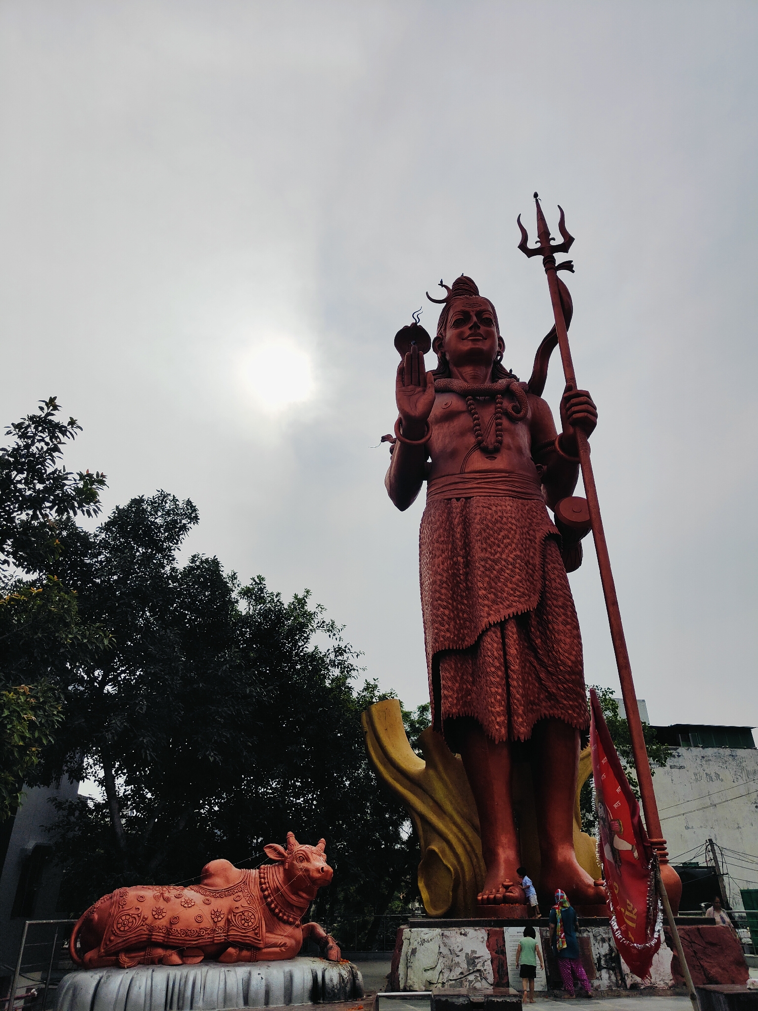 The image depicts a large, reddish-brown statue of the Hindu deity Shiva, standing tall against a hazy, overcast sky. The statue is intricately detailed, showing Shiva holding a trident in his right hand and a flame in his left. He wears a patterned skirt and several necklaces. To the left of Shiva, a statue of a reclining bull, Nandi, is visible, adorned with ornate patterns. In the foreground, a few individuals, including two children and an adult, are interacting with the base of the Shiva statue, with one child appearing to touch its foot. A red flag with text is planted near the statue's base. The background includes lush green trees and a white building. The scene is located at the Sheetla Mata Mandir in Gurgaon, India.