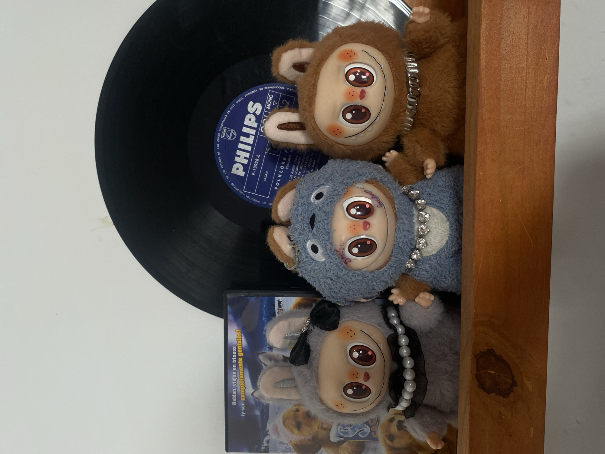 A vinyl record, a DVD case, and three stuffed animals are arranged in a wooden box. The record is black with a blue Philips label, bearing the text "FOLKLORE." The DVD case has a visible title, "Hablan, viajan en trineos," with the text "y son completamente geniales!" underneath. The three stuffed animals are monkey-like creatures with large, expressive eyes. The top animal is brown, the middle one is blue, and the bottom one is grey. They are adorned with accessories such as chains and bows. The wooden box has a simple, rectangular construction. This scene is in Buenos Aires, Argentina, as indicated by the SADAIC label on the record, which is an Argentinian collecting society.