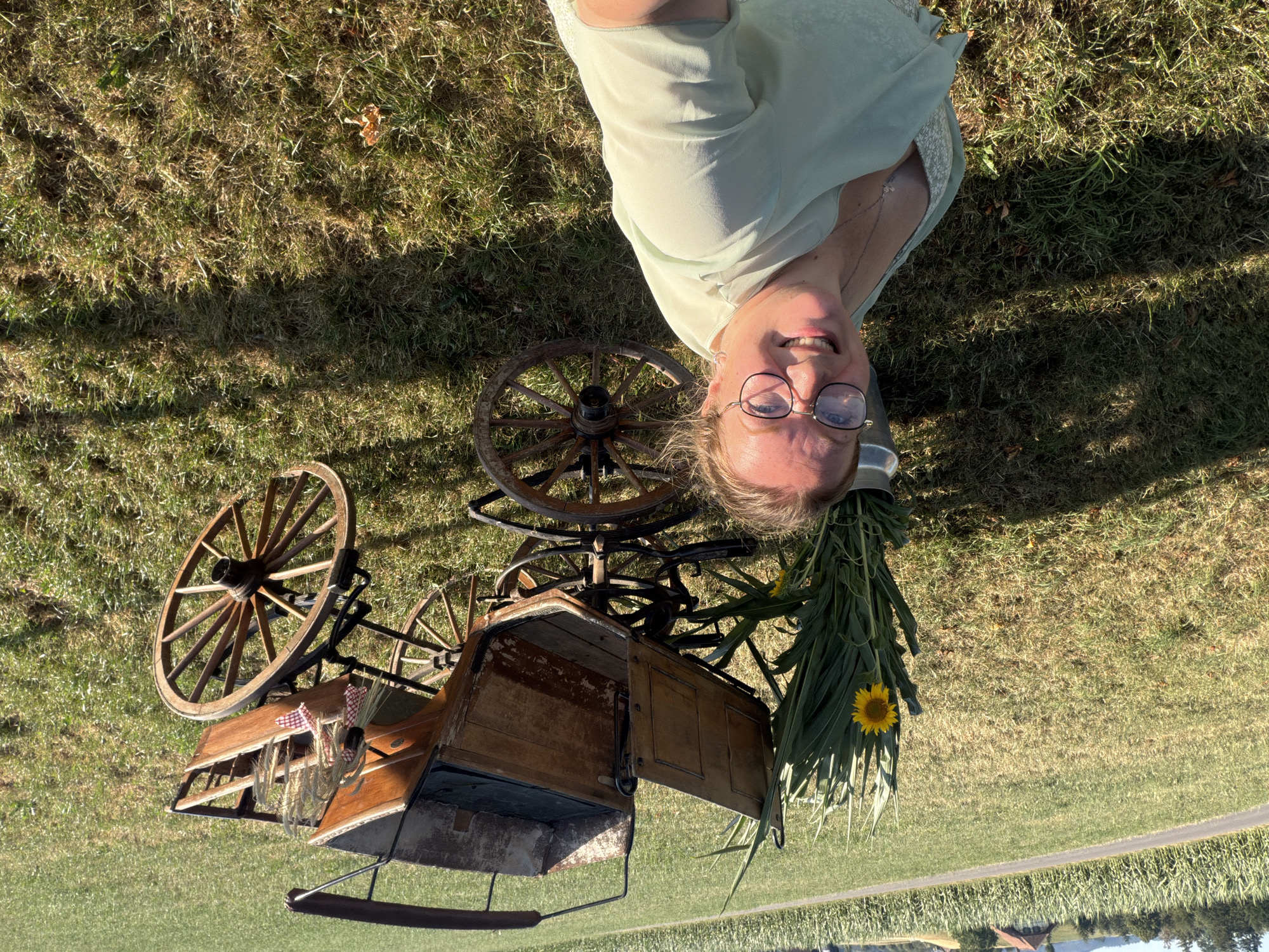 A woman is taking a selfie outdoors in a grassy field. She is wearing glasses and a light green dress with a floral pattern. Behind her, to her left, is a bucket with a bouquet of sunflowers and tall green stalks. To her right is an antique wooden carriage with large spoked wheels. The carriage has a dark upholstered seat and a weathered wooden exterior. A small checkered ribbon is tied to some stalks of wheat, which are placed in the back of the carriage. In the background, a cornfield stretches towards a rural landscape with trees and buildings. The sun is casting shadows on the grass. The image was taken in Ufhusen, Switzerland.