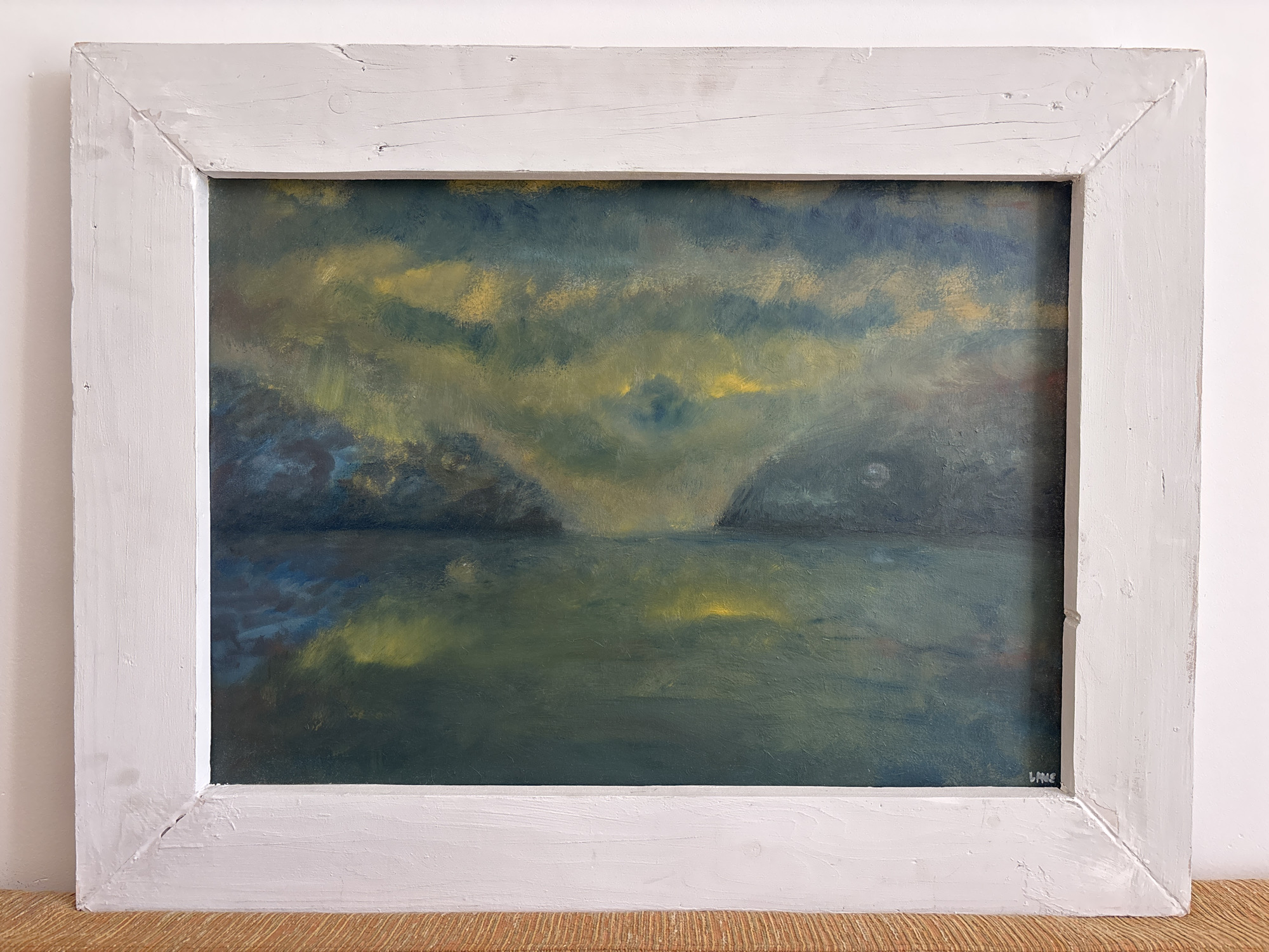 A framed oil painting depicts a twilight seascape with mountainous landforms on either side. The sky is filled with dramatic, swirling clouds in shades of dark blue, gray, and yellow. A pale, hazy light emanates from the center of the composition, suggesting a setting or rising sun partially obscured by clouds. The water in the foreground reflects the colors of the sky and the land, with streaks of yellow and green. The landmasses are rendered in dark blues and greens, with textured brushstrokes. The painting is housed in a distressed, white wooden frame. The signature "LANE" is visible in the bottom right corner of the painting. The painting is displayed on a surface that appears to be a woven mat or textured fabric, with a plain white wall in the background. The painting is not located in the city of Bečići, Montenegro.