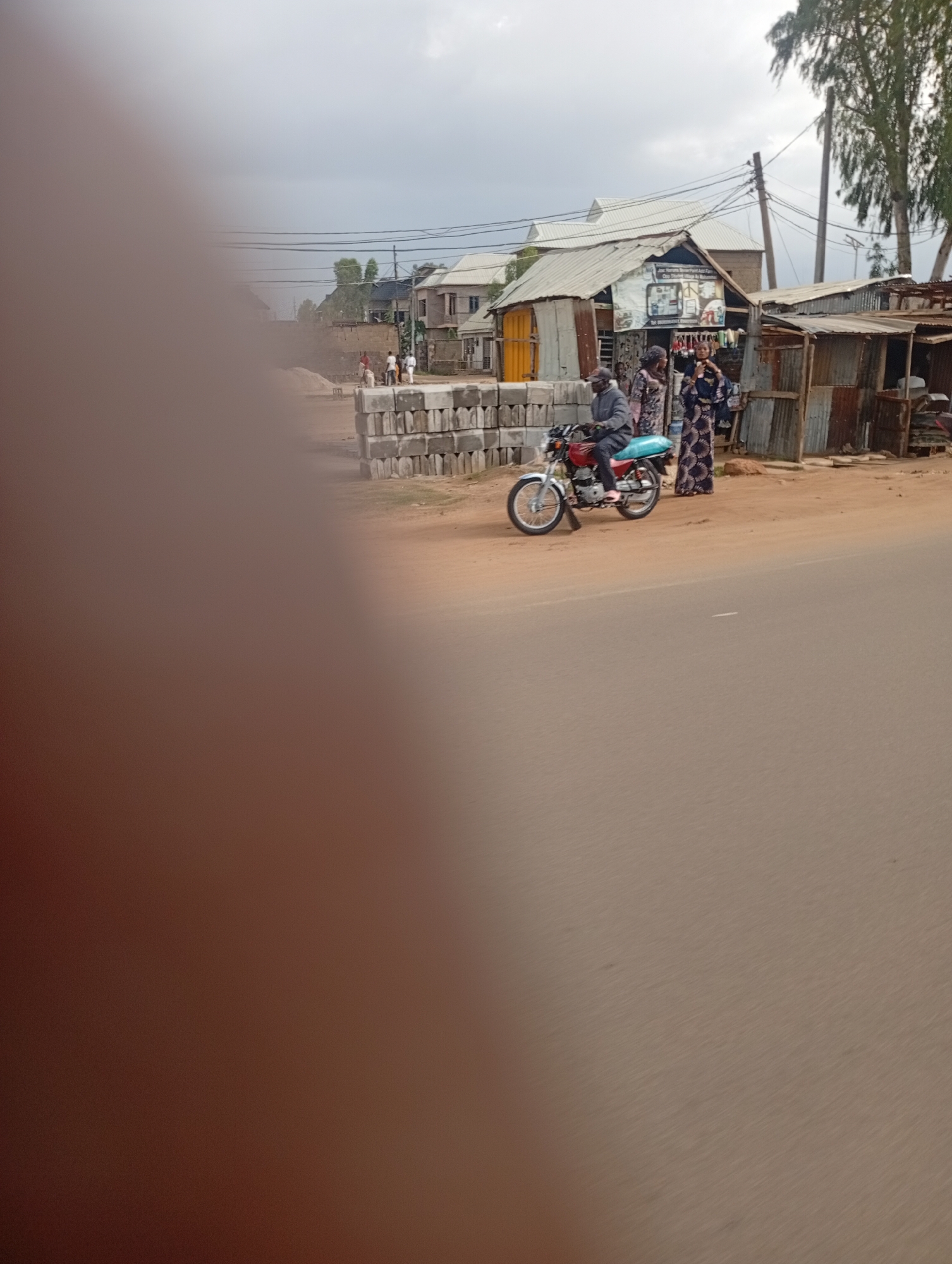 A man is riding a red motorcycle in an outdoor scene. He is wearing a grey jacket and is facing to the right. There is a light blue bag on the back of the motorcycle. Two women are standing near the motorcycle. One woman is wearing a dark blue and white patterned dress, and the other is wearing a pink and black patterned dress. They are looking at each other. In the background, there are several buildings with corrugated metal roofs. There are also stacks of grey concrete blocks on the ground. The scene appears to be a street in a town or village in Jos, Nigeria. The road is made of dirt, and there are power lines visible in the sky. The overall lighting suggests it is daytime, with an overcast sky.