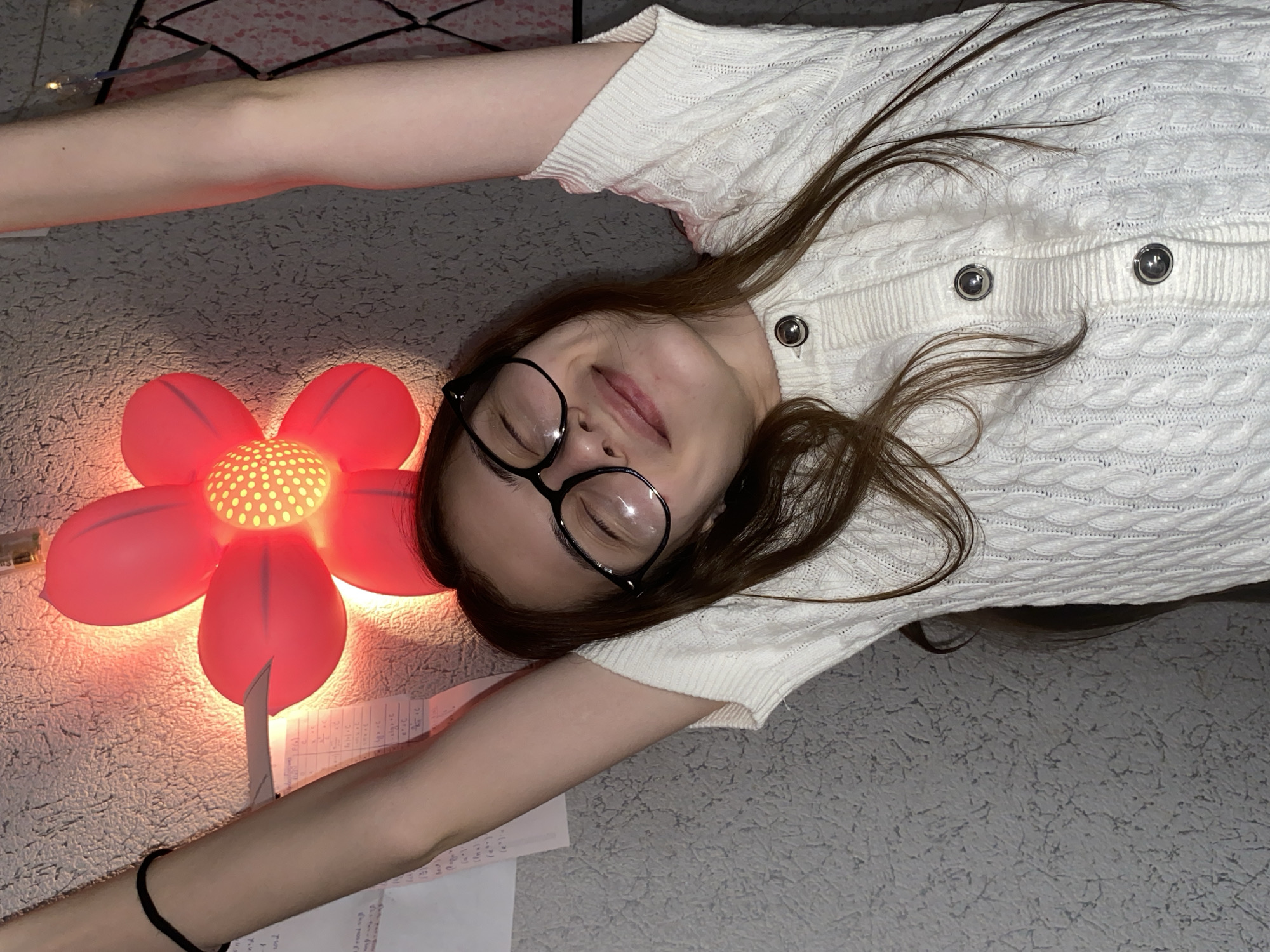 A woman is lying on a patterned surface next to a red flower-shaped lamp with a lit center. She is wearing glasses and a white cable-knit sweater with buttons. Papers are scattered near her outstretched arm. The location in Votkinsk, Russia, cannot be determined from the image.