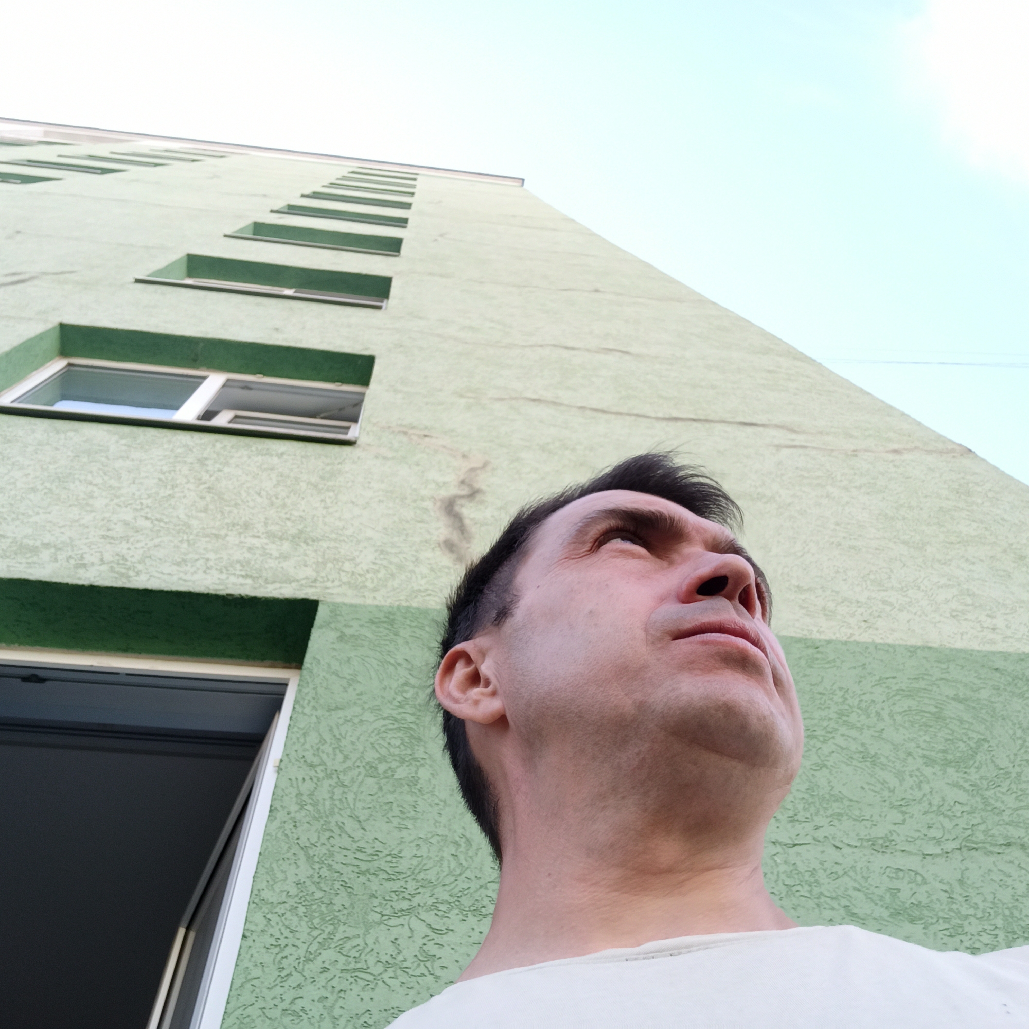 An extreme low-angle, full shot captures a man looking upwards, partially obscuring the pale green, stucco-textured facade of a multistory apartment building. The building features multiple rows of horizontally elongated, recessed windows, some of which are visible and appear to be open. The sky above is a pale blue. The man, wearing a light-colored t-shirt, is positioned in the foreground, with his face turned to the right and slightly upwards. His hair is dark and short, and he has a visible ear and the side of his neck. The image's perspective emphasizes the scale of the building in relation to the man. The photograph was taken in Nizhny Novgorod, Russia.