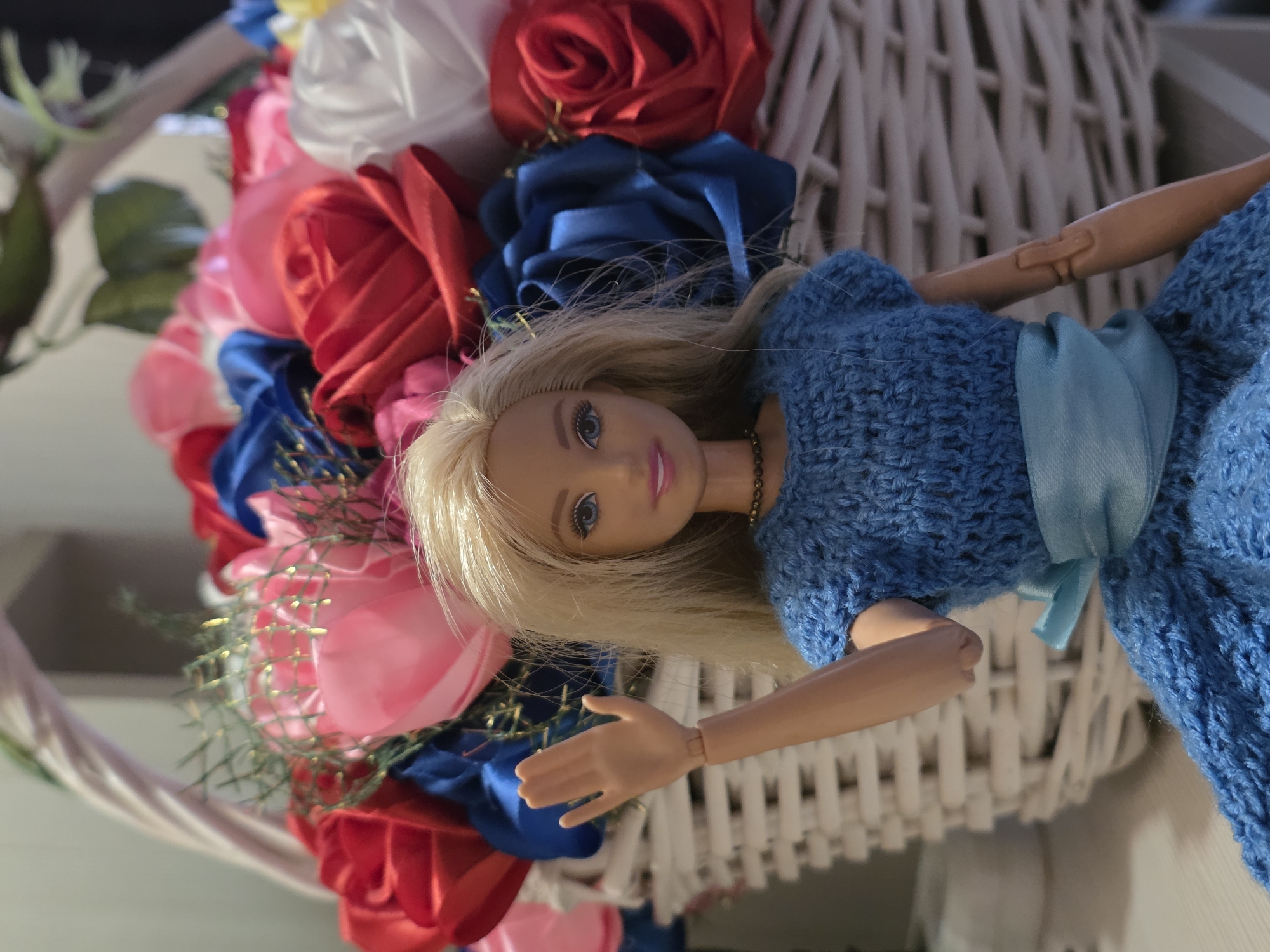 A blonde doll with blue eyes and pink lips, wearing a blue knitted dress and a blue ribbon belt, is standing in front of a white woven basket filled with red, pink, blue, and white satin roses. The doll is waving with its right hand, palm facing forward. The background is blurred, showing hints of a light-colored wall and possibly green foliage. The scene is brightly lit, with the focus on the doll and the flowers.
