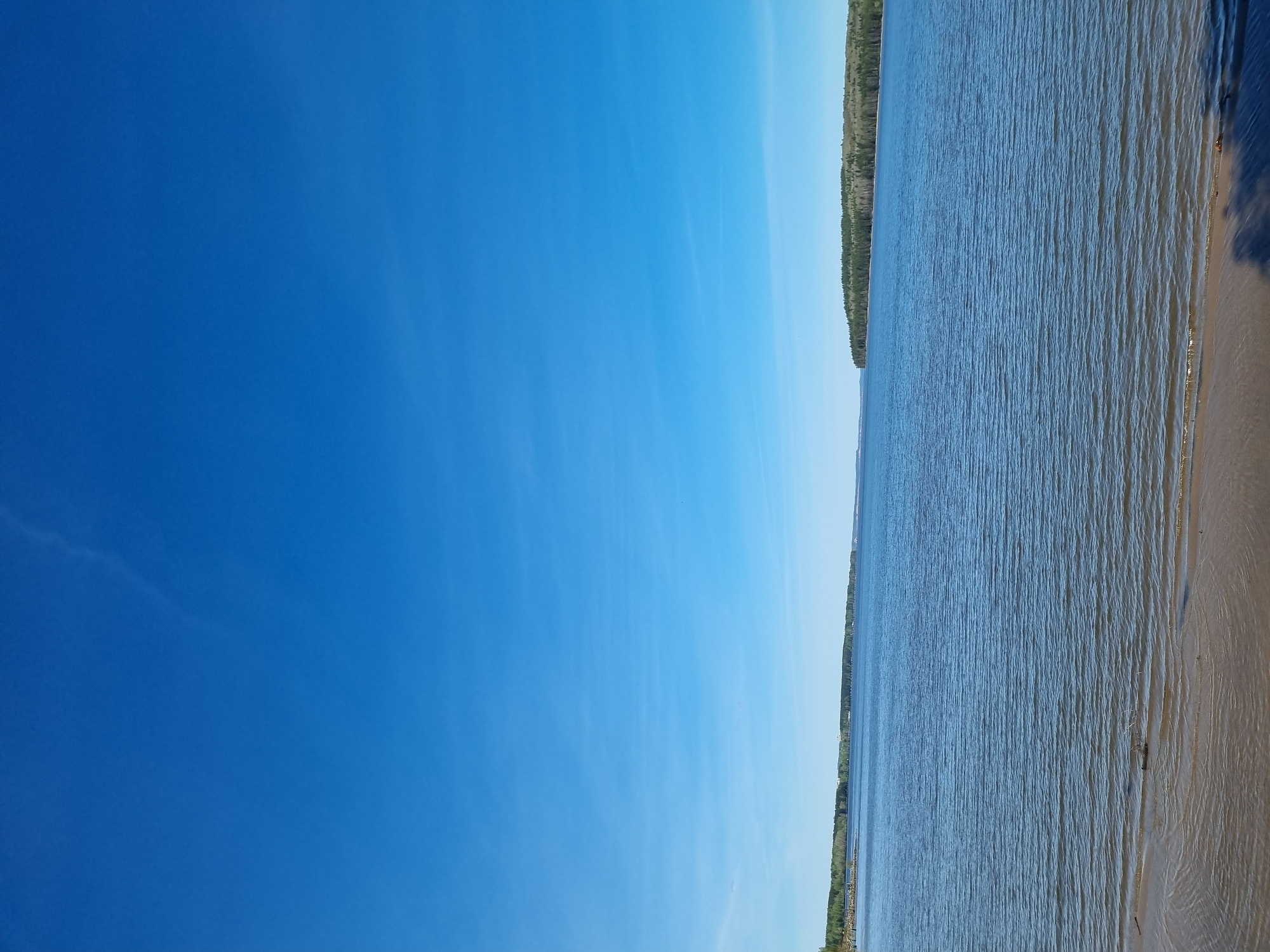 A bright blue sky takes up most of the image on the left. To the right is a body of water, possibly the Kama River, with a thin line of trees along the opposite shore. The edge of a sandy beach is visible on the far right. The image is oriented vertically, suggesting it was taken with a mobile device. Considering the information given, the scene is likely somewhere in Chaikovsky, Russia, as the Kama River is a notable feature of that city.